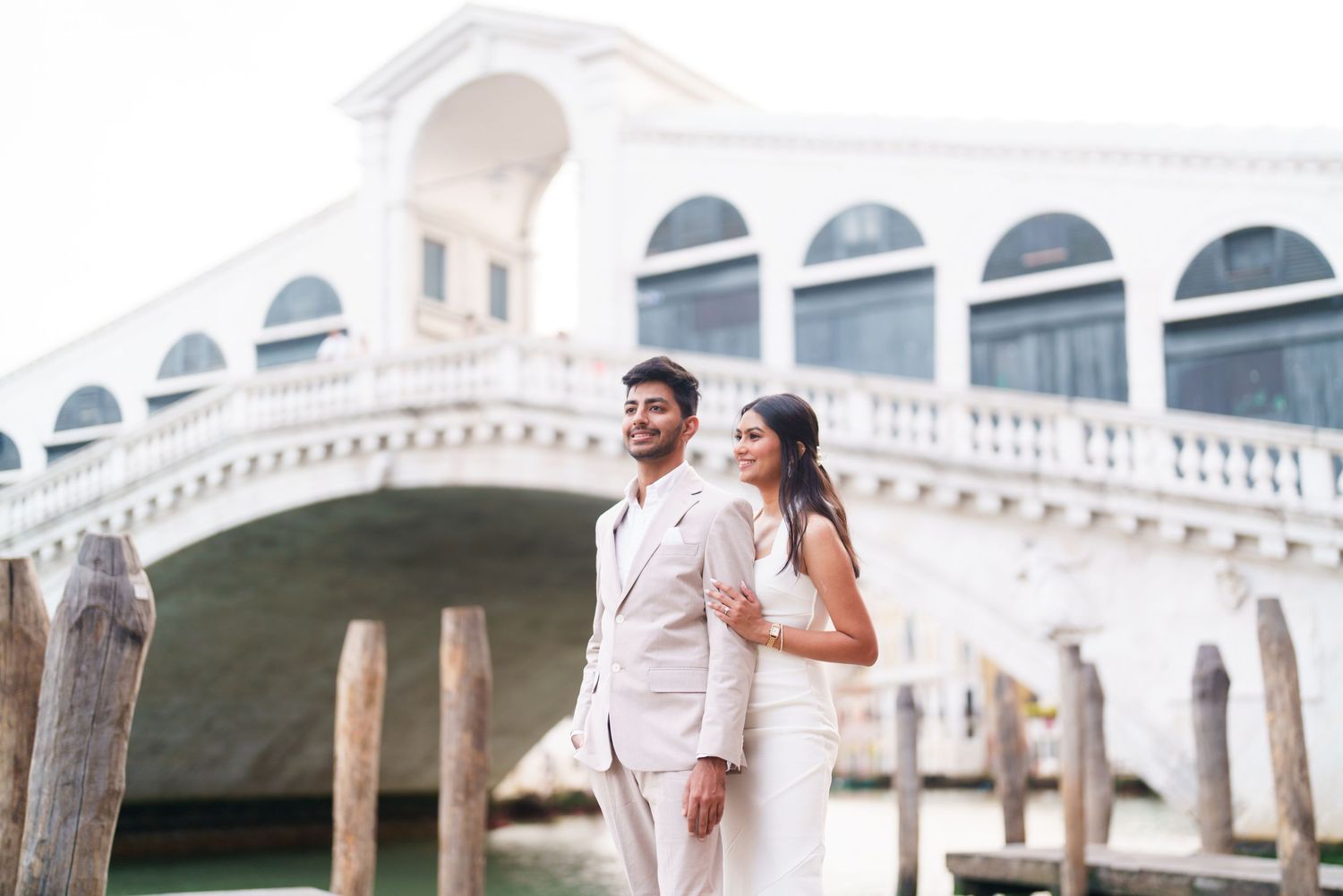 A couple in white wedding attire walks along a grand ornate balcony with arched architectural details.