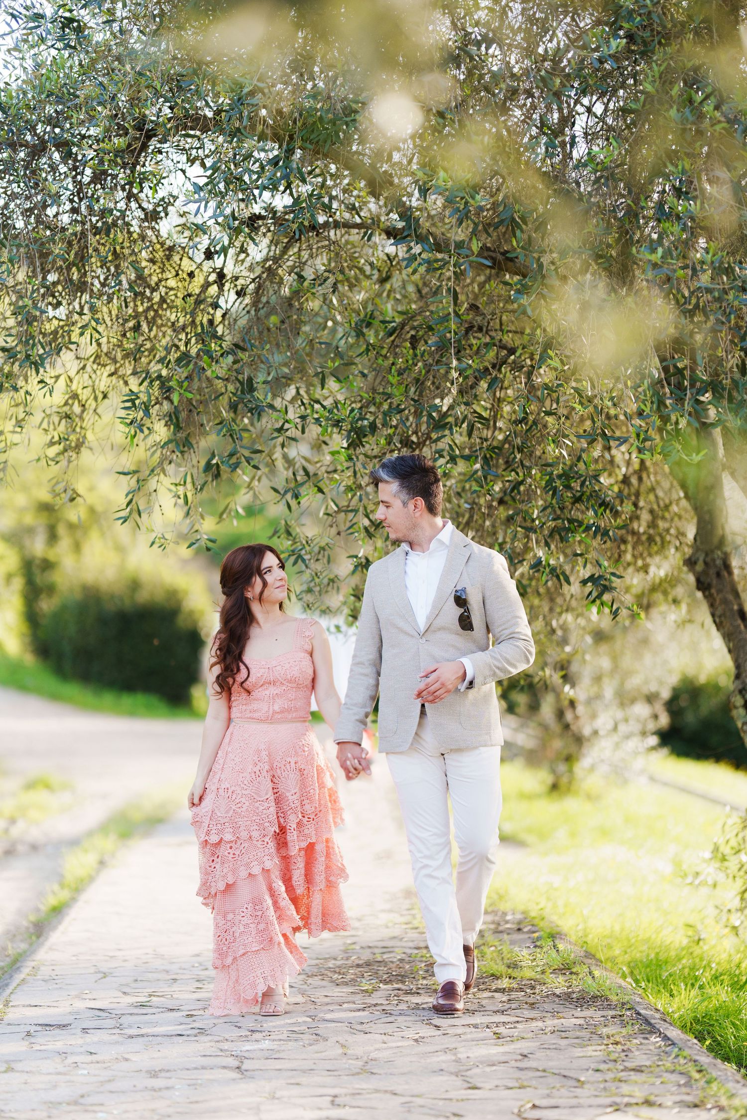 A couple in pink and white attire walks hand in hand down a stone path surrounded by olive trees in golden sunlight.