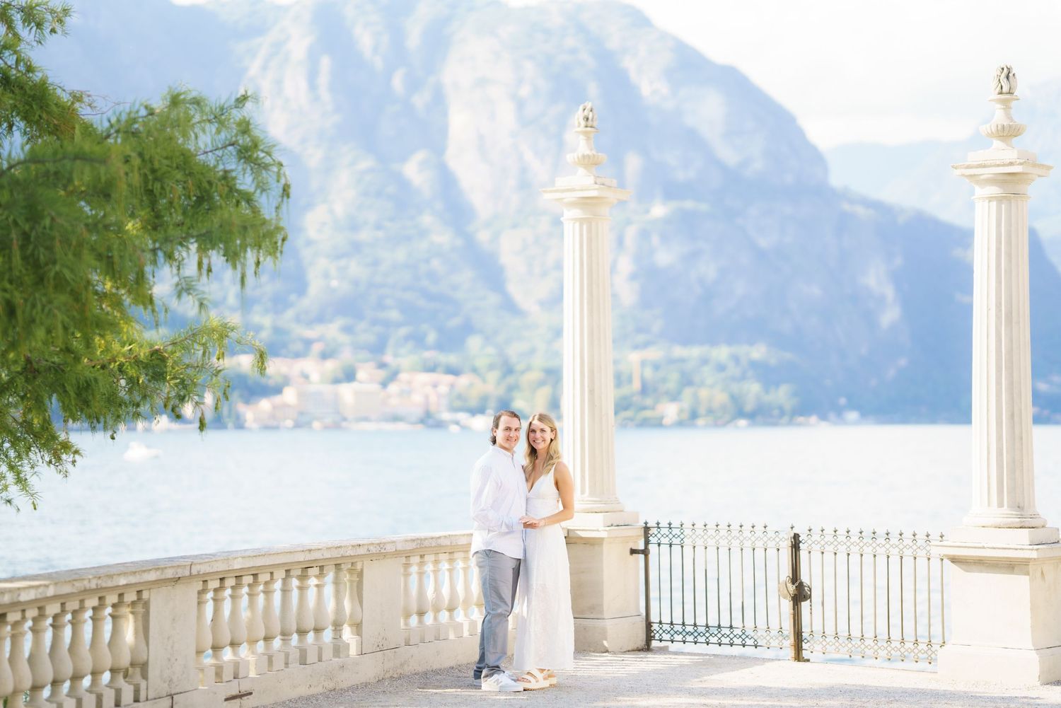 A romantic couple stands on an elegant white balcony overlooking a scenic mountain lake vista on a bright sunny day.
