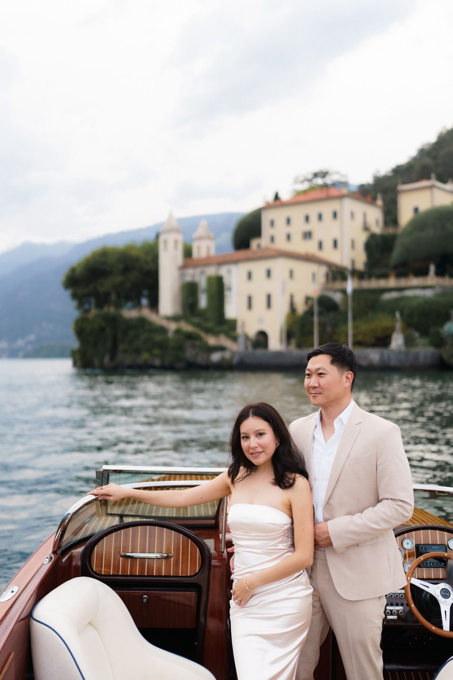A couple in wedding attire poses on a classic wooden boat with Lake Como and Italian villas in the background.