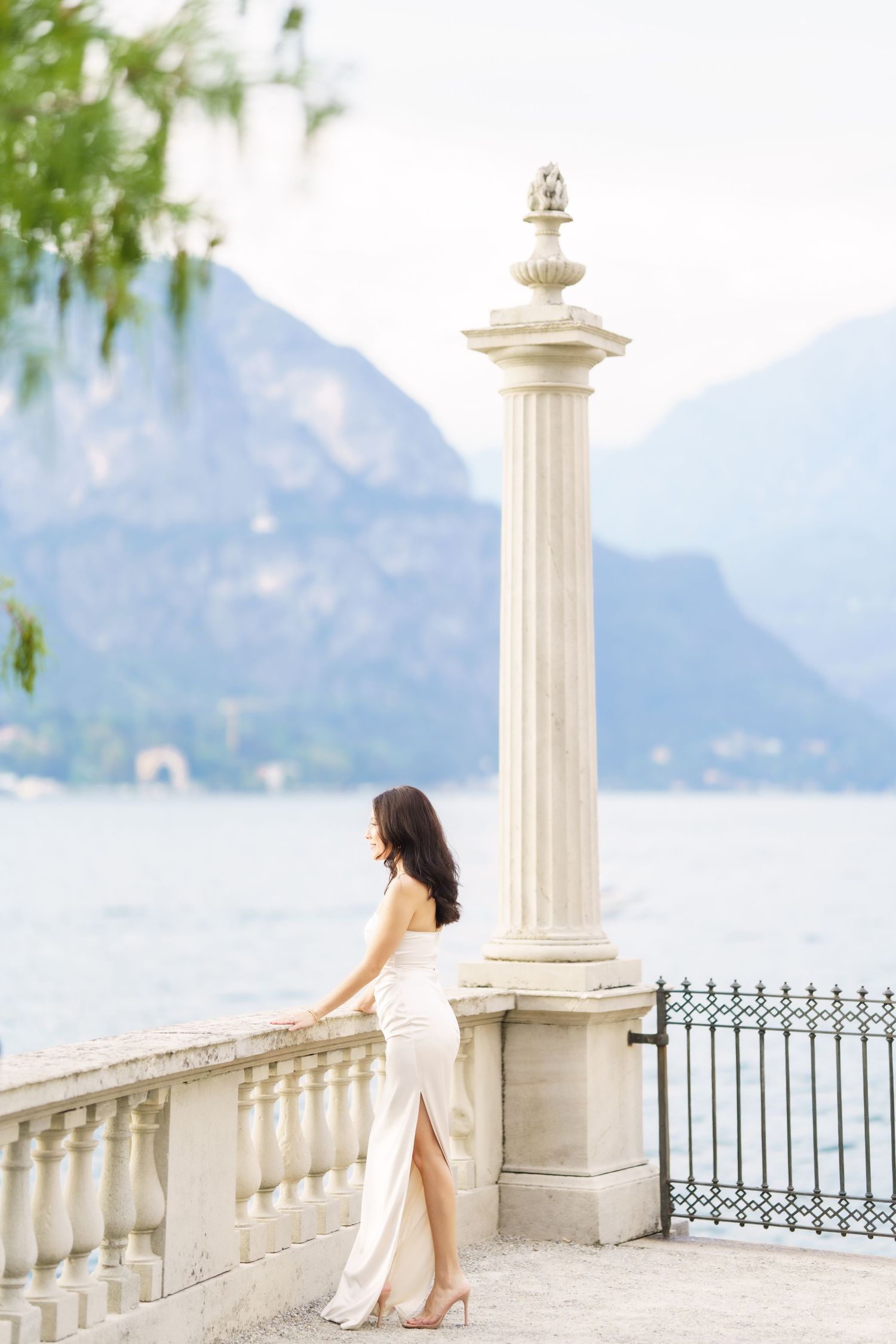 Elegant white dress silhouette against ornate stone column and railing overlooking scenic mountain lake landscape.