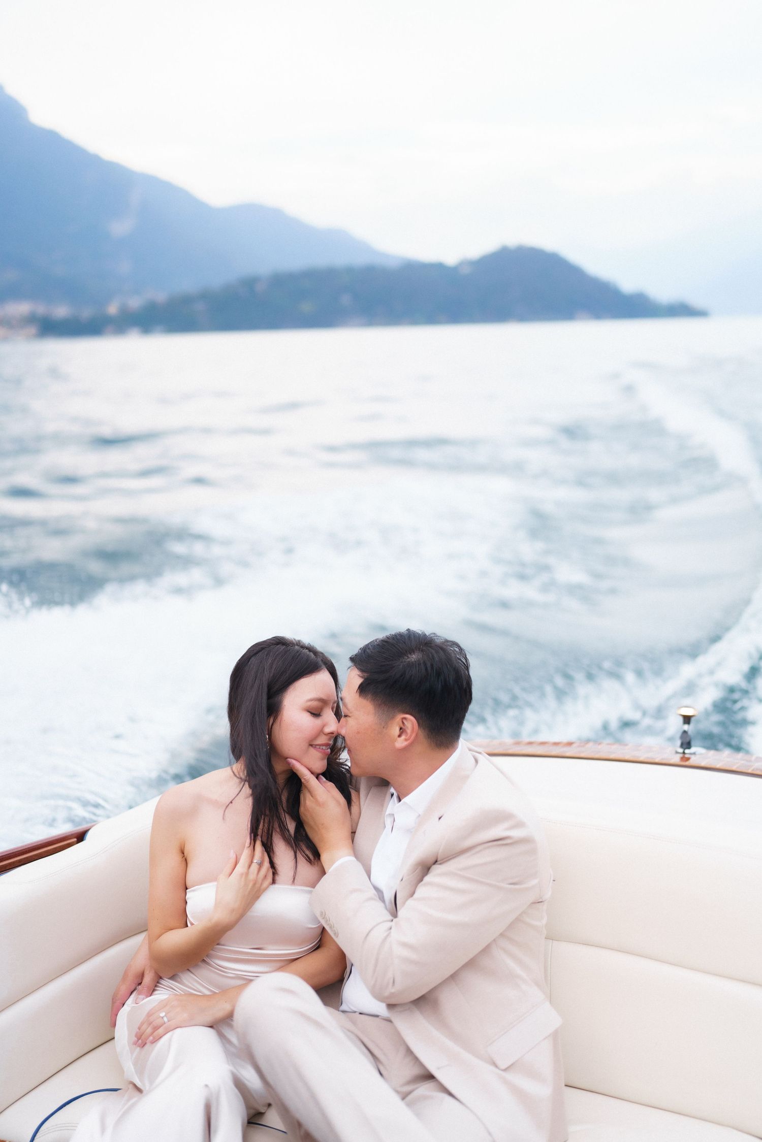 Romantic couple in light colored outfits sharing an intimate moment on a luxury boat with mountains in background.