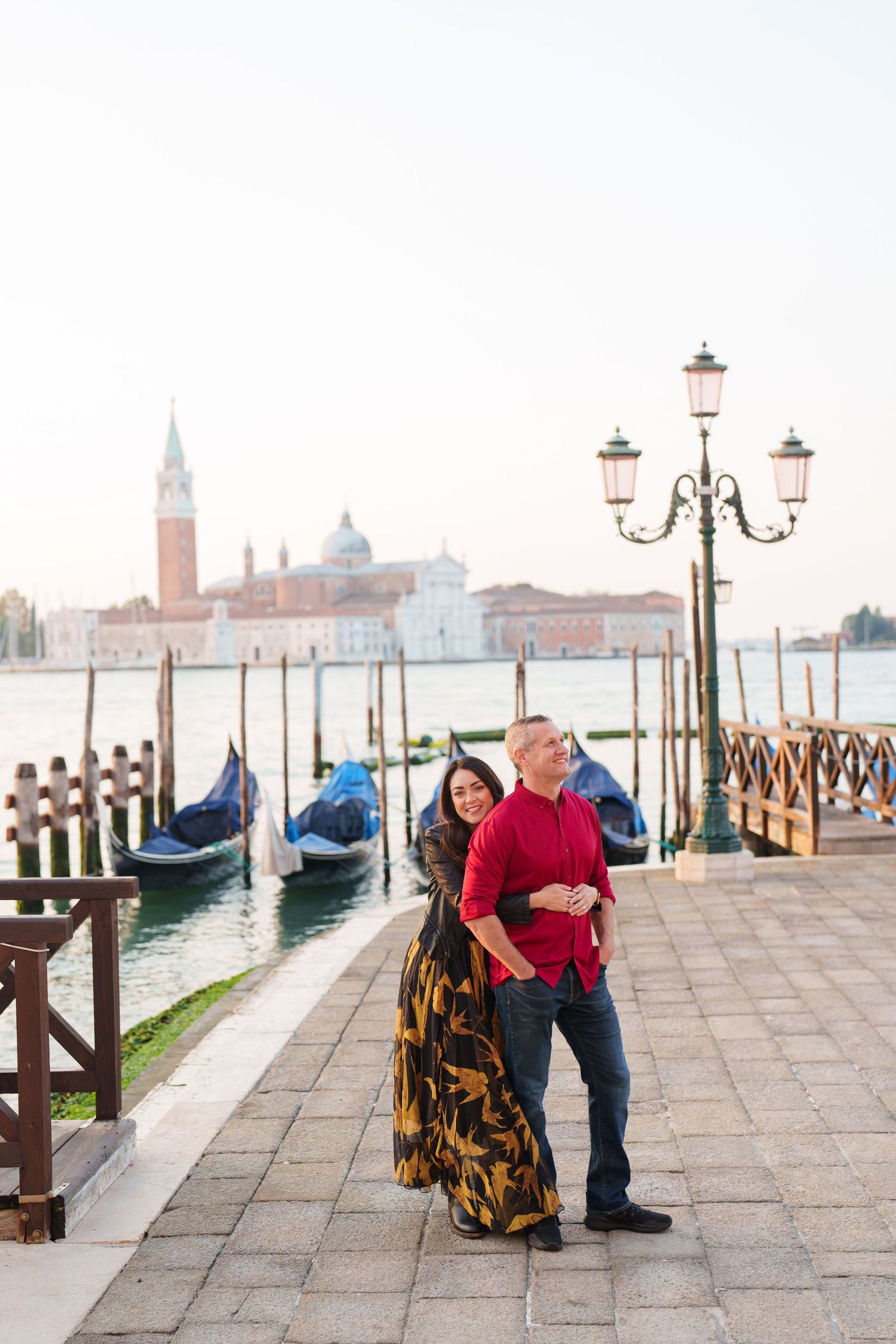Romantic couple poses along Venice's iconic waterfront with gondolas and San Giorgio Maggiore church in the background.