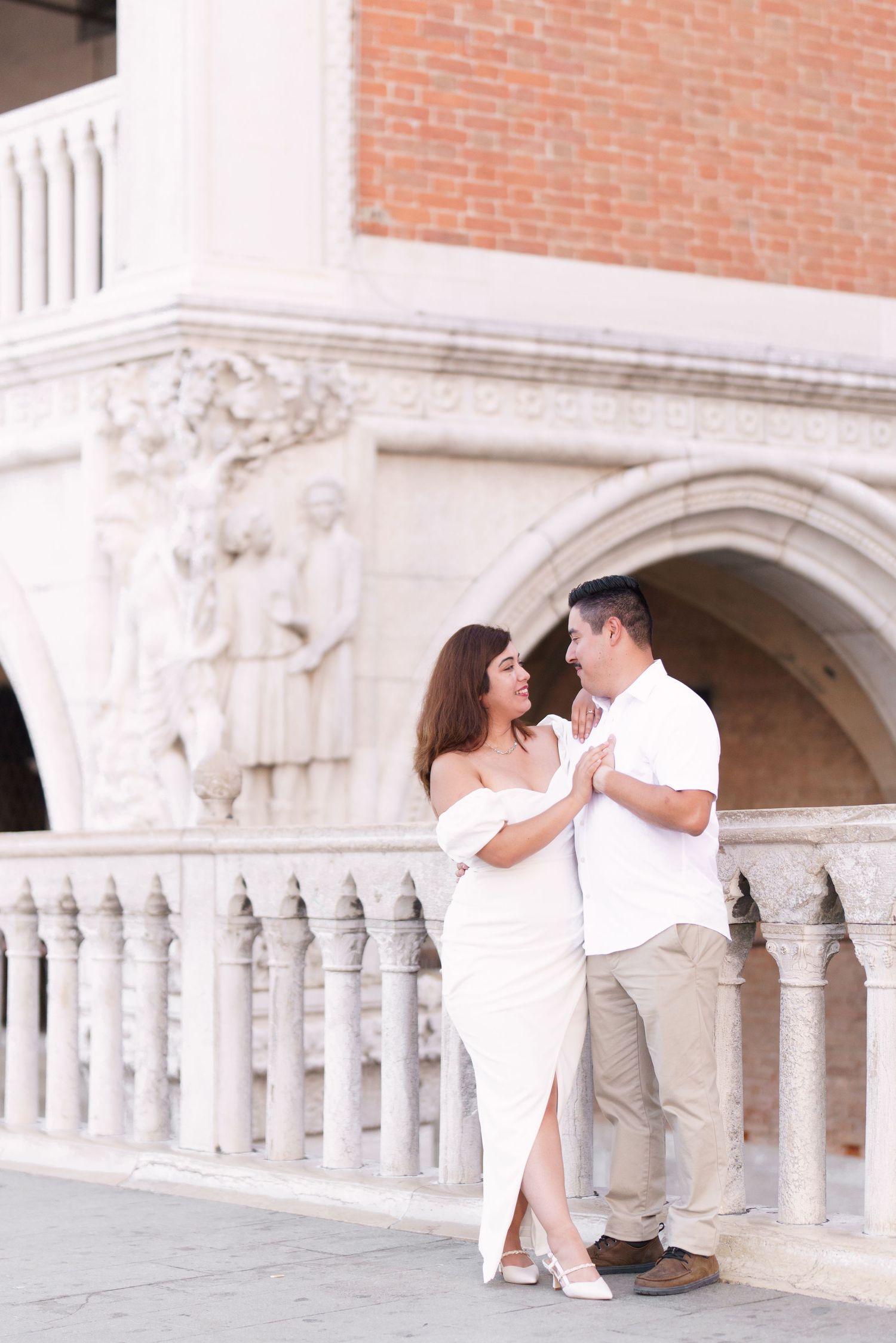 Romantic couple in white outfits embracing by ornate stone columns and brick architecture in Venice, Italy.