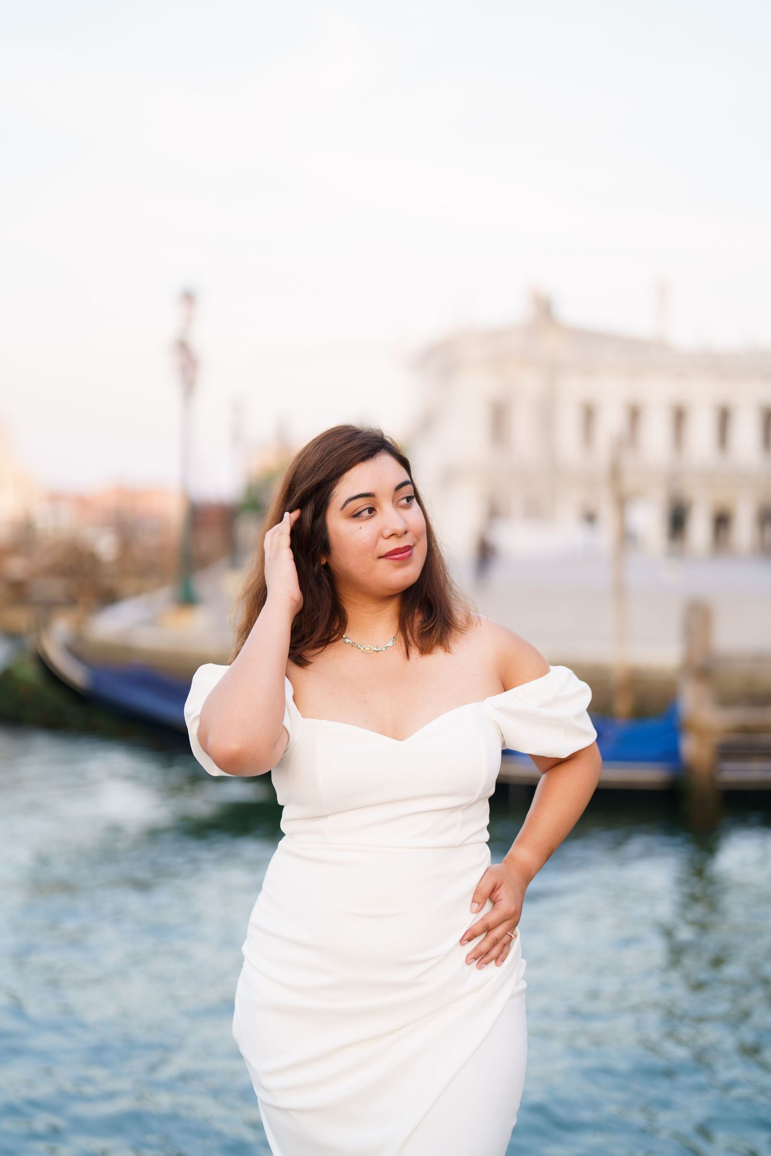 A person in a white off-shoulder dress poses elegantly by the Venice canals at sunset.
