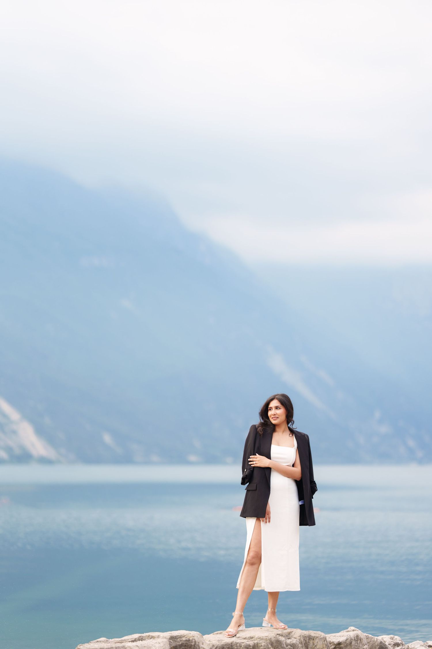 A person in a white dress and black jacket stands on coastal rocks with misty mountains and ocean in the background.