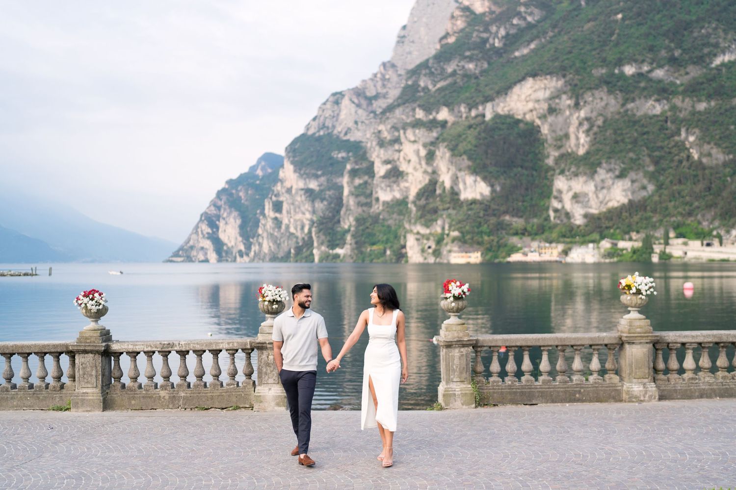 A couple holds hands while walking along a scenic lakeside promenade with mountain cliffs in the background.