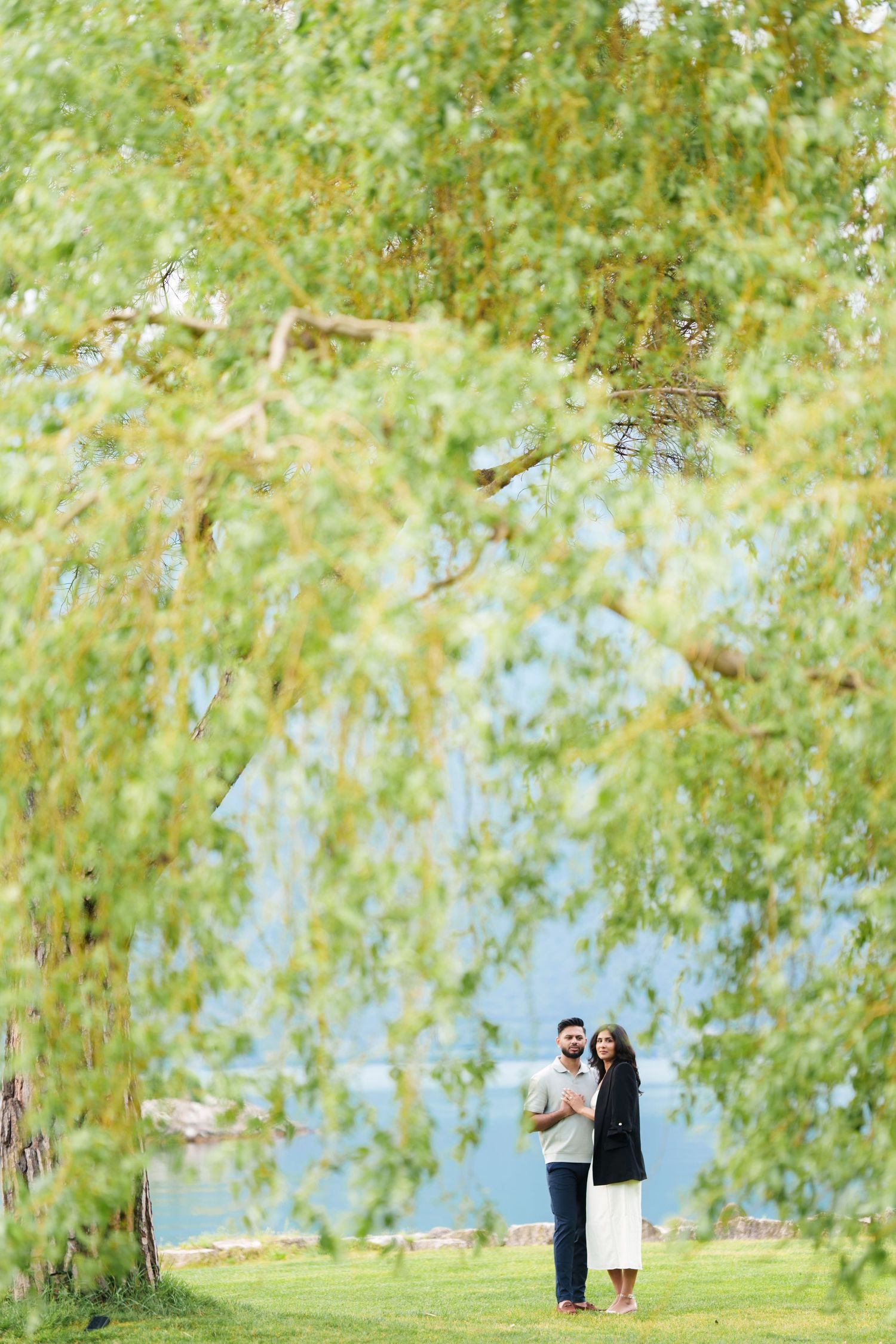 A couple stands together under drooping willow tree branches on a grassy lawn near water on a sunny day.