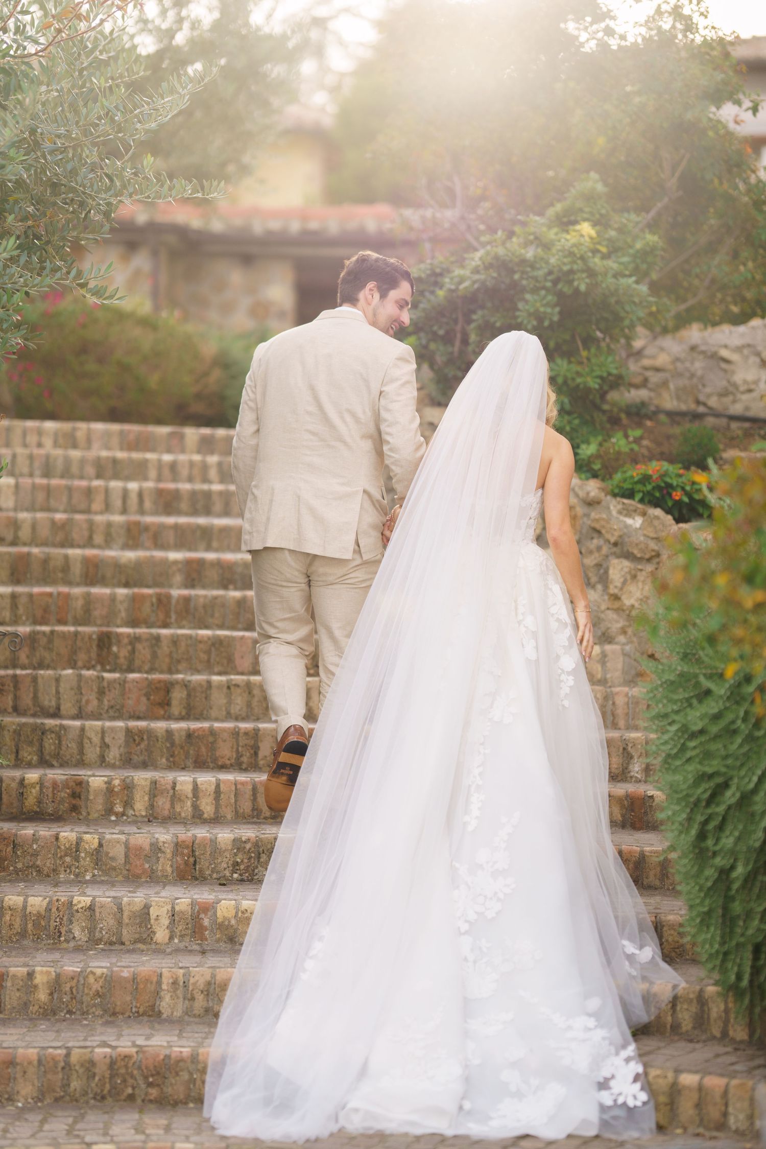 Romantic wedding couple ascending stone garden steps with flowing white dress trailing behind.
