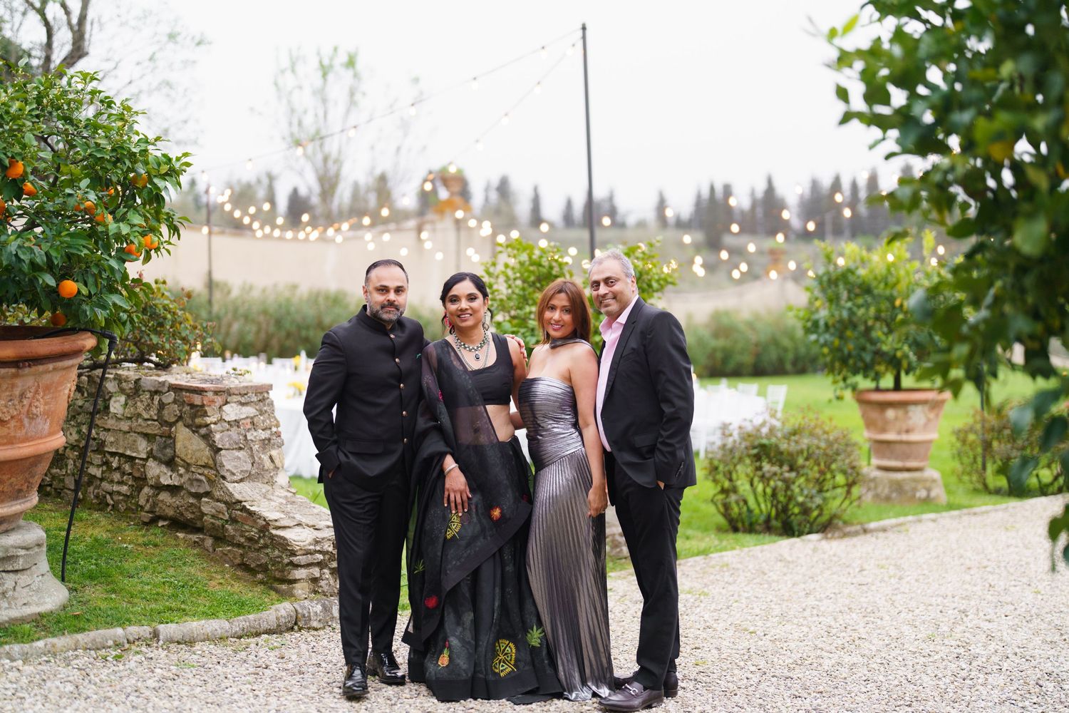 Group photo of wedding guests in formal attire standing on a gravel path with stone wall and garden backdrop.