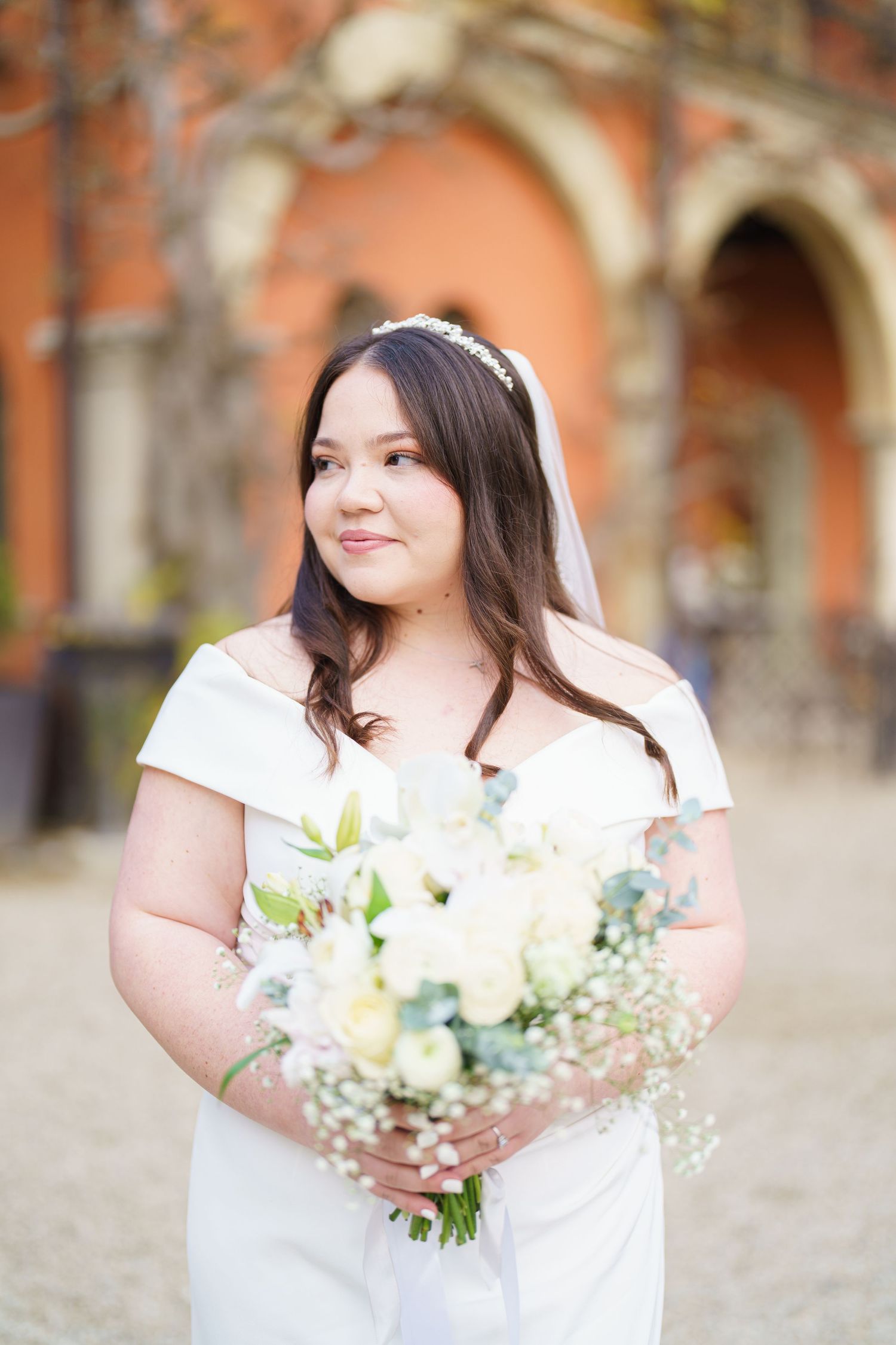 A bride in a white dress holds a bouquet of white flowers against a peach-colored building backdrop.