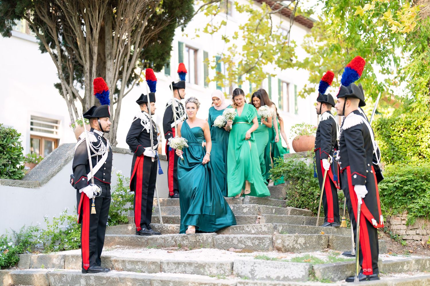 Military officers in dress uniforms and wedding party members on stone garden steps.