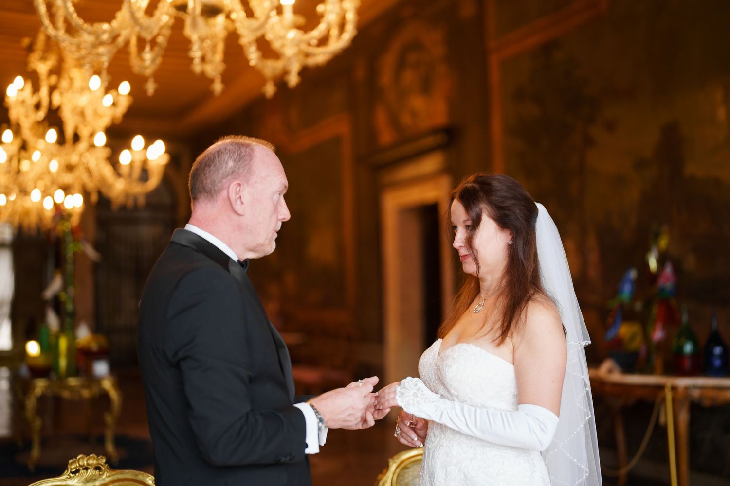 A couple shares their first dance as newlyweds in an elegant ballroom with ornate chandeliers overhead.