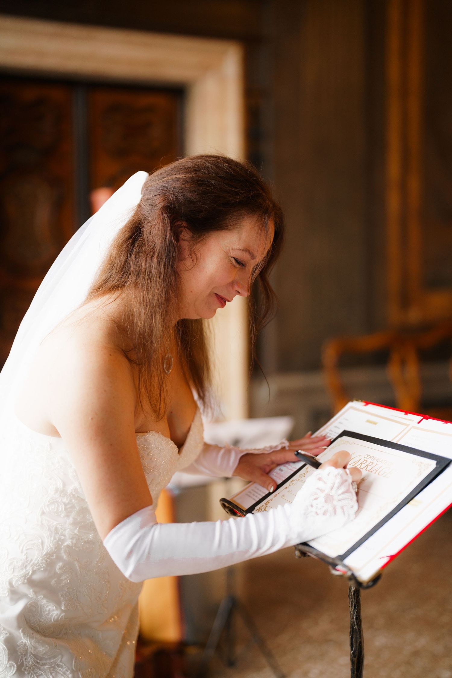 A bride in a white wedding dress and long gloves smiles while signing a document during her ceremony.