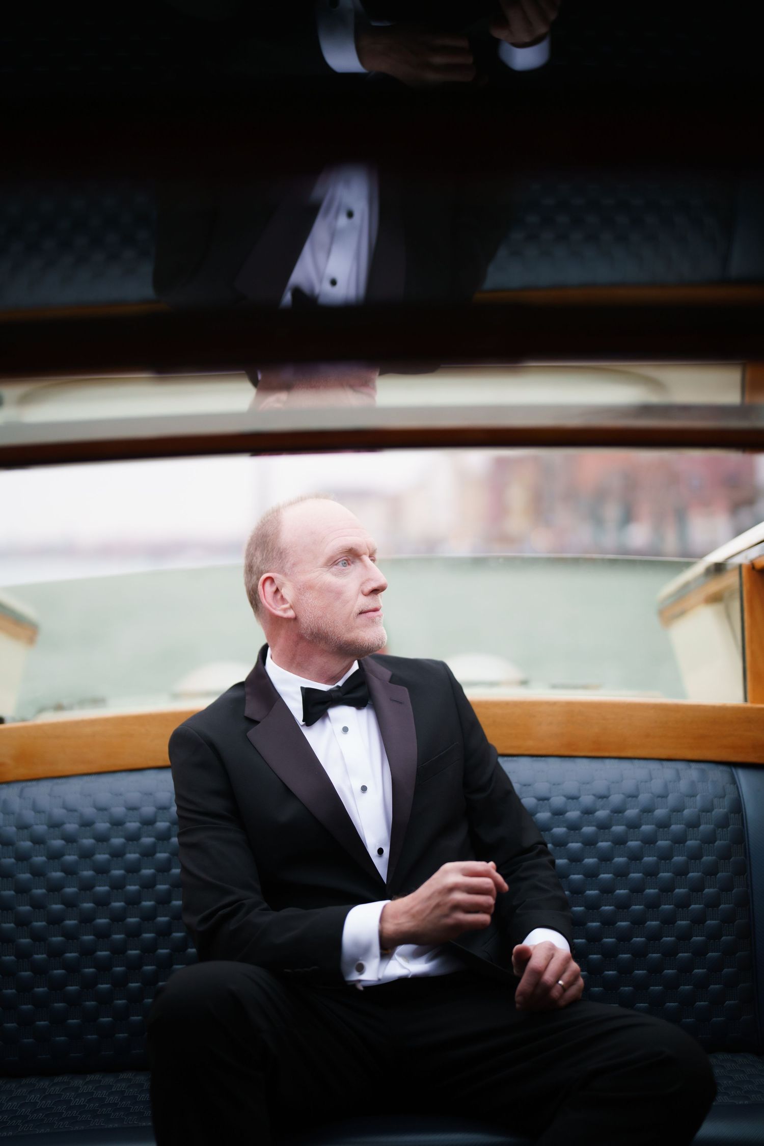 Person in formal black tuxedo sits on dark upholstered couch looking to the side.