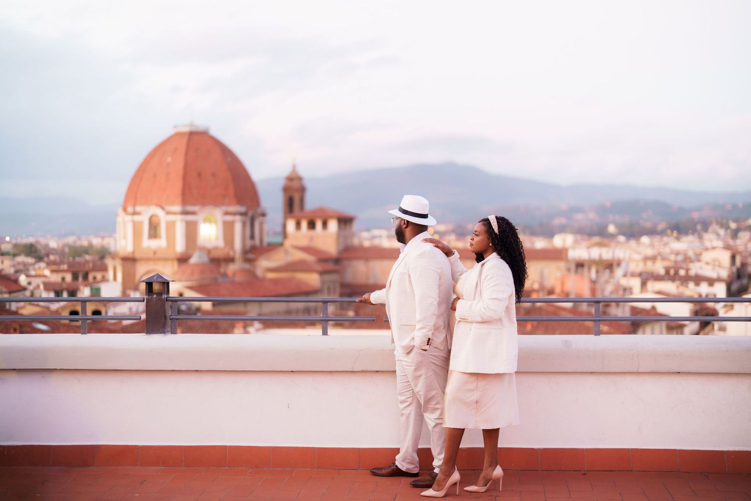 Couple in white outfits standing on rooftop terrace with Florence Cathedral dome and city skyline backdrop.