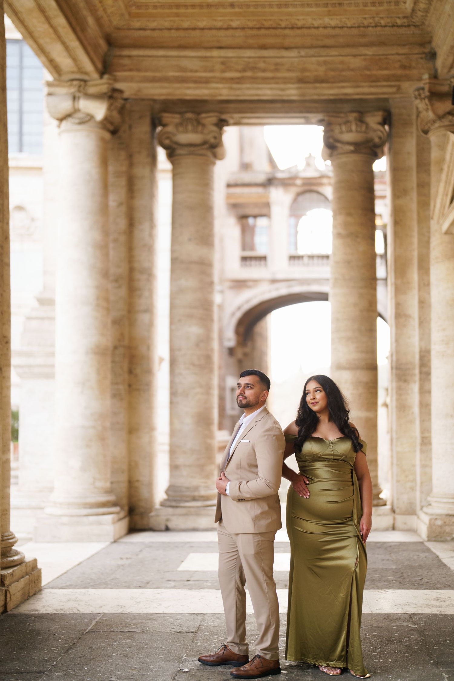 Elegant couple posing among grand stone columns in Rome, woman in olive green satin gown, man in beige suit.