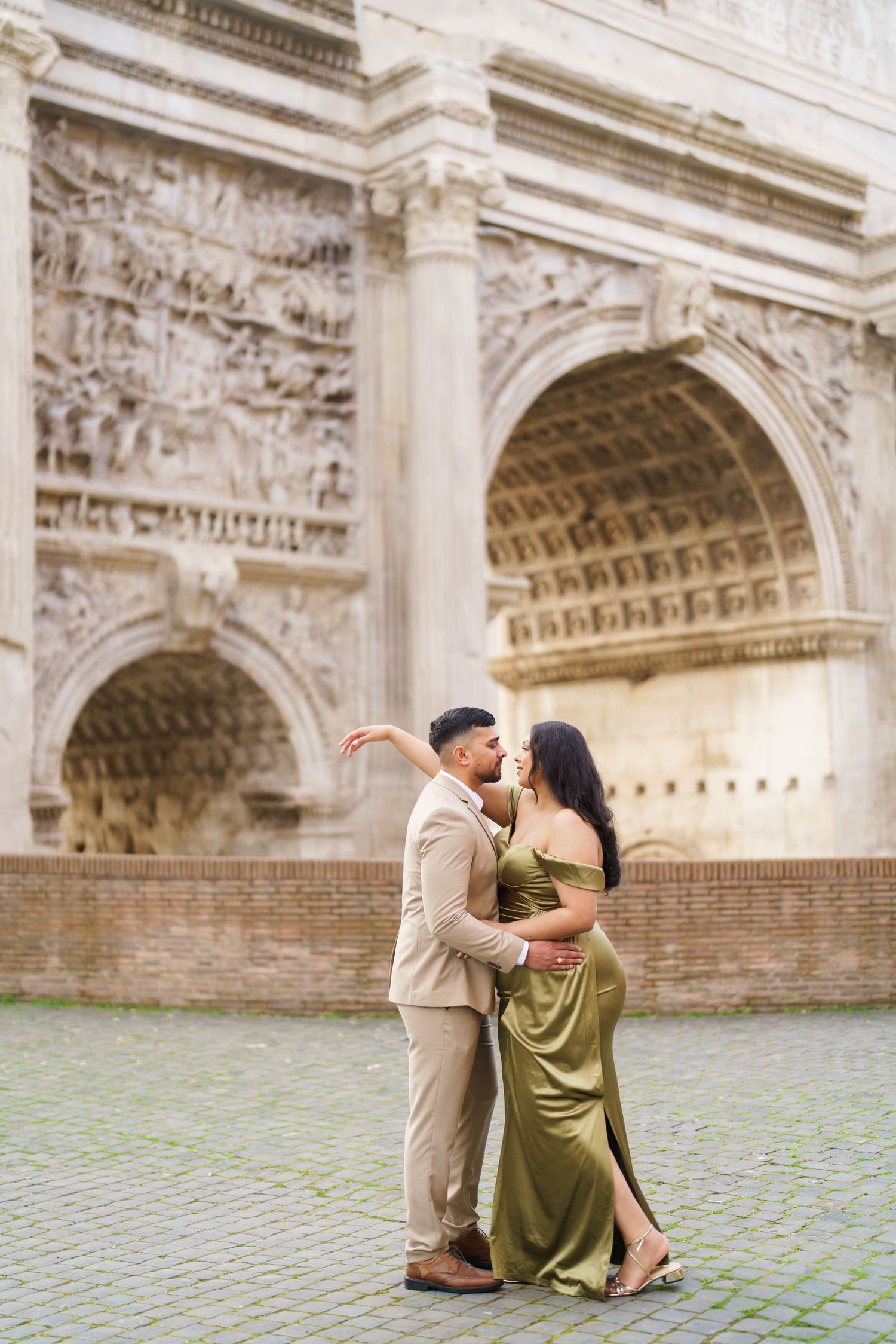Couple embracing in front of the Arch of Constantine in Rome, woman in green gown, man in beige suit.