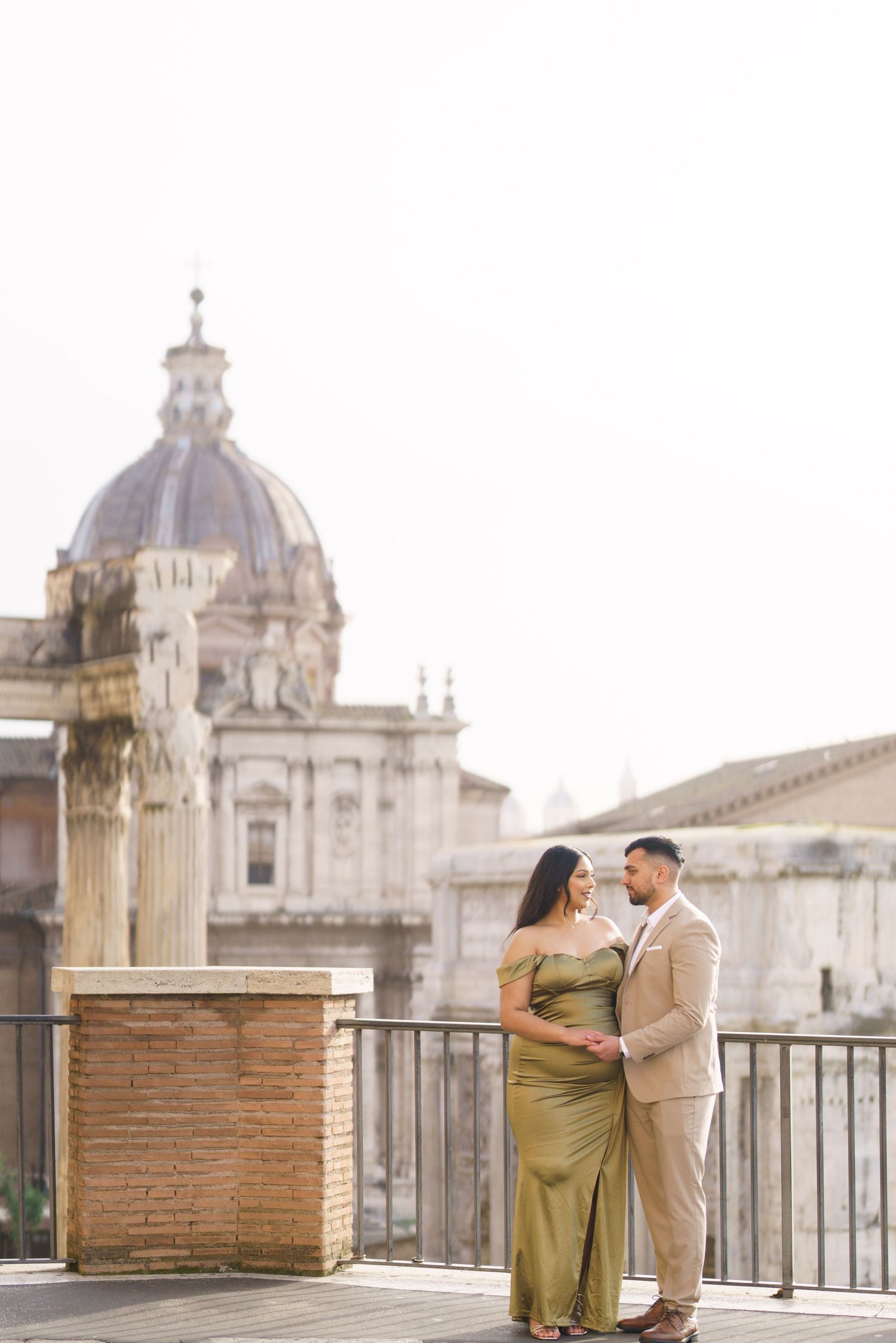 Couple in elegant attire poses on a terrace with Rome's historic domed architecture in the soft background.