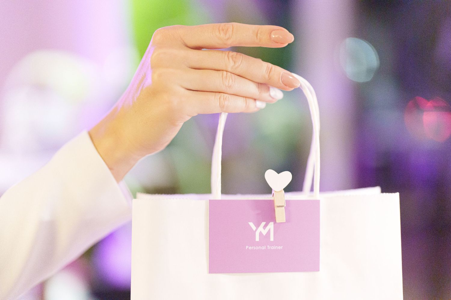 A hand holding a white gift bag with a pink YM logo label against a soft bokeh background.