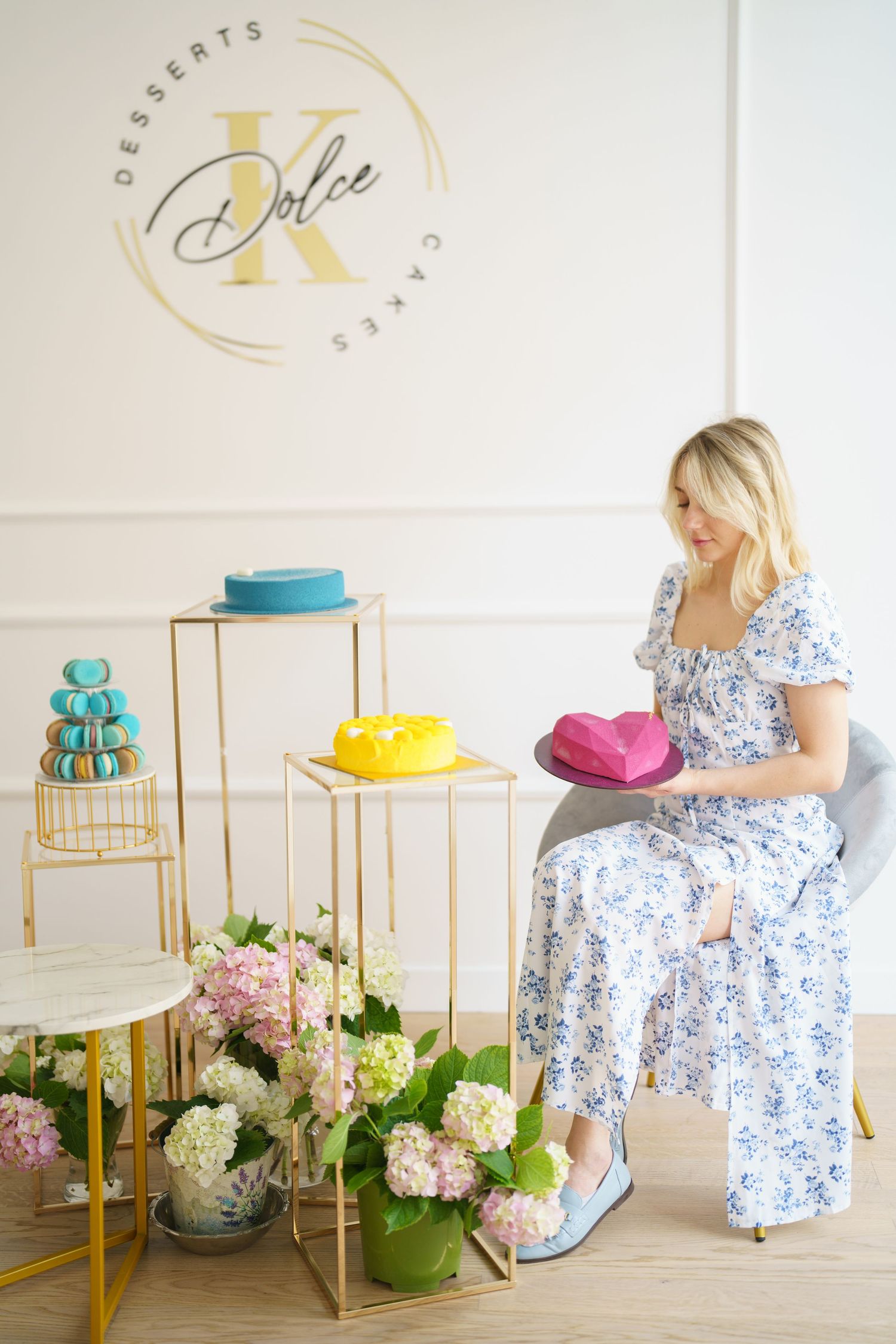 Blonde woman in floral dress holding pink cake at Dolce K desserts shop with colorful cakes and hydrangeas.