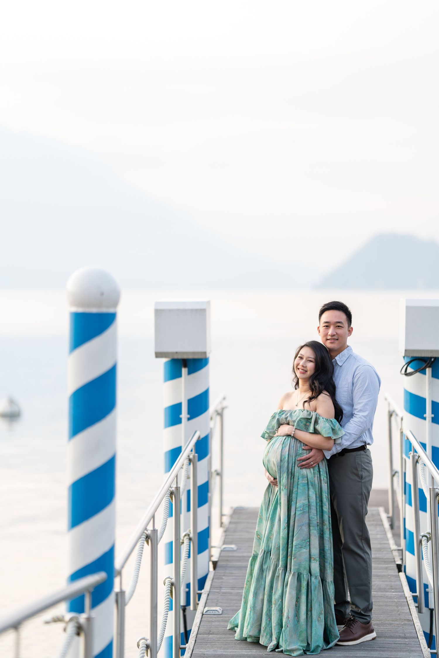 Couple poses on a waterfront pier, woman in green maternity dress embraced by man in blue shirt with mountains behind.
