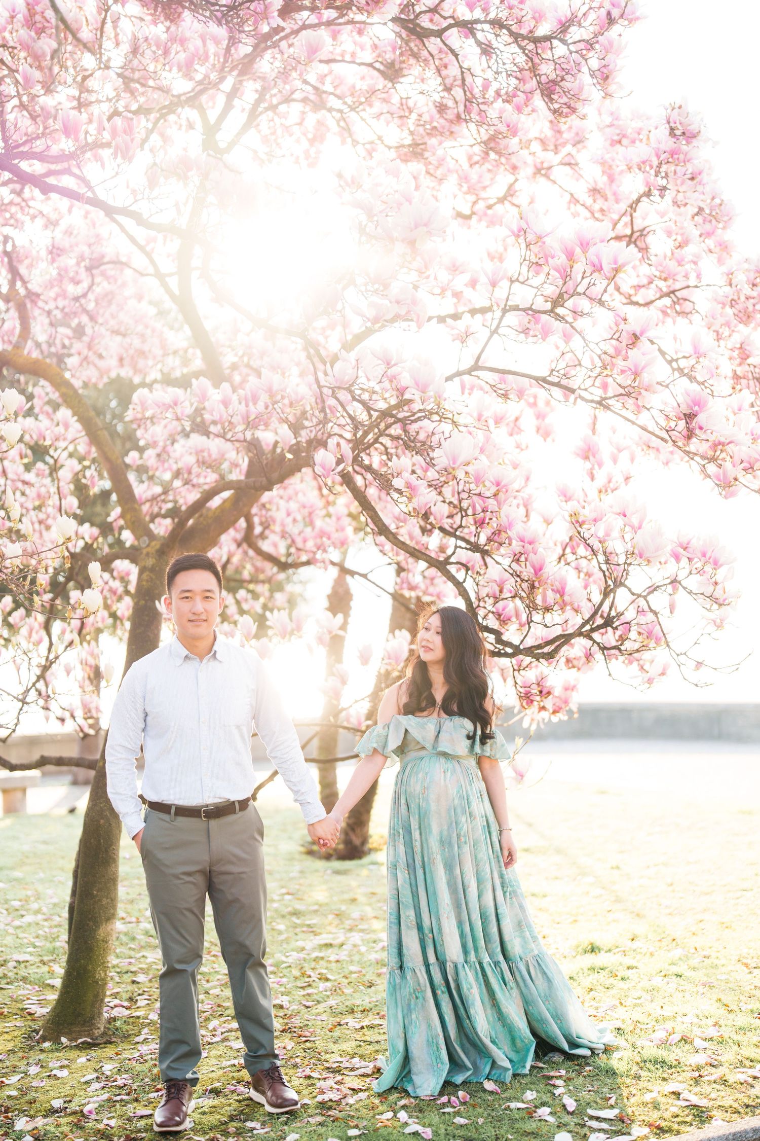 Couple holding hands under blooming pink magnolia tree, woman in teal floral dress, man in white shirt, soft golden light.