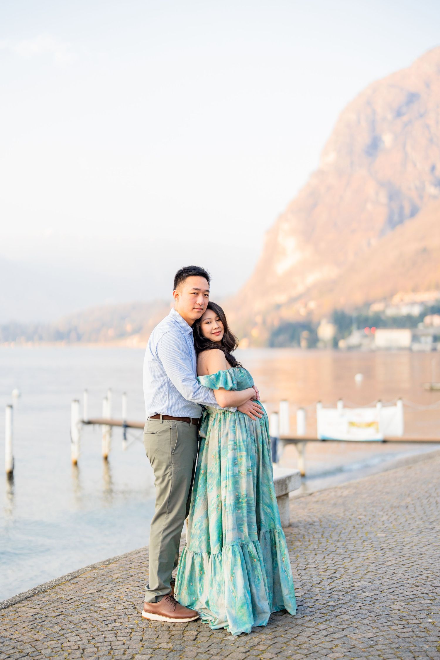 Couple embracing by a lakeside promenade with mountain backdrop, woman in teal maxi dress, golden hour light.