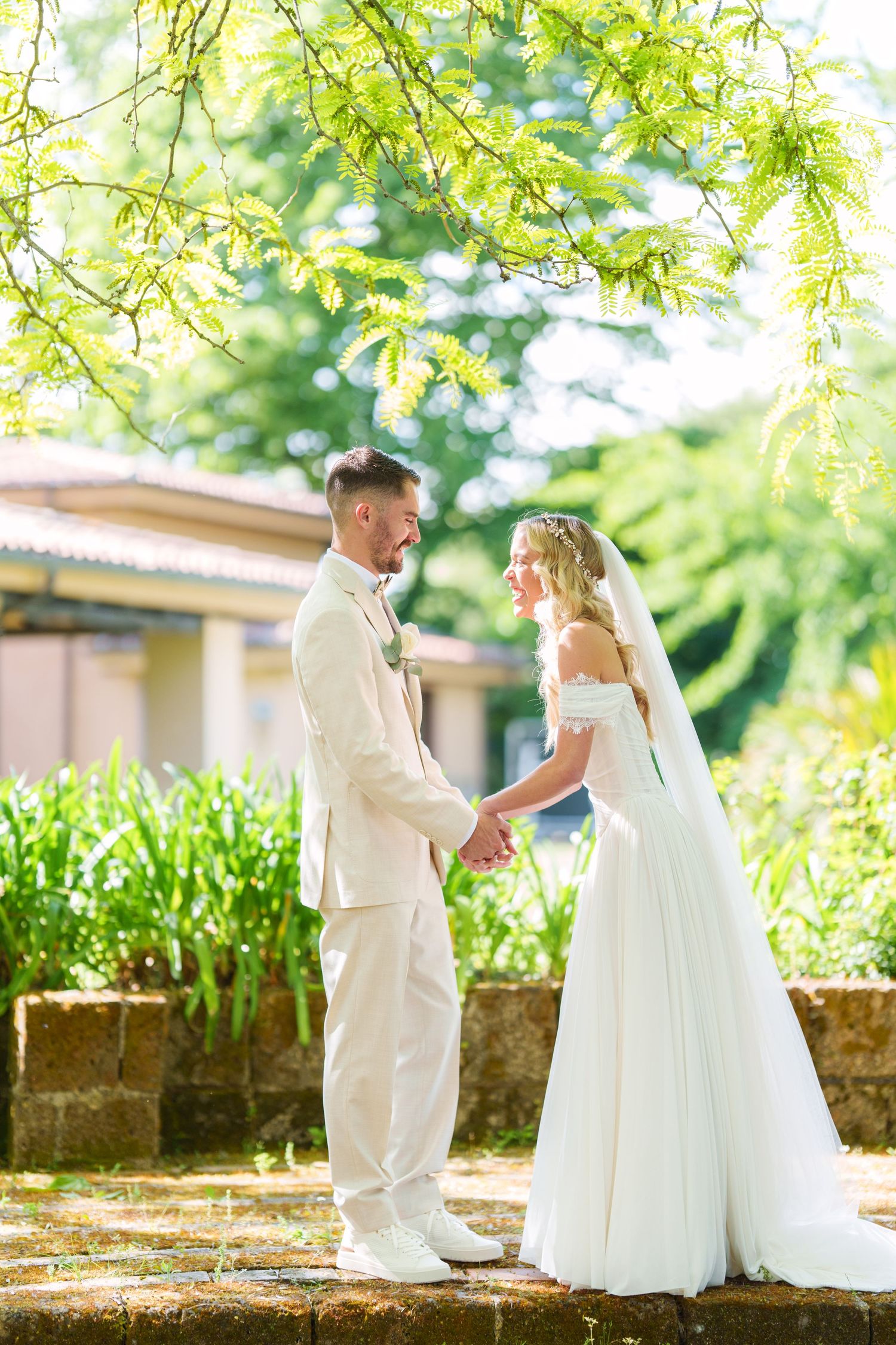 Bride and groom hold hands in a sunlit garden, wearing white wedding attire, surrounded by lush green trees.