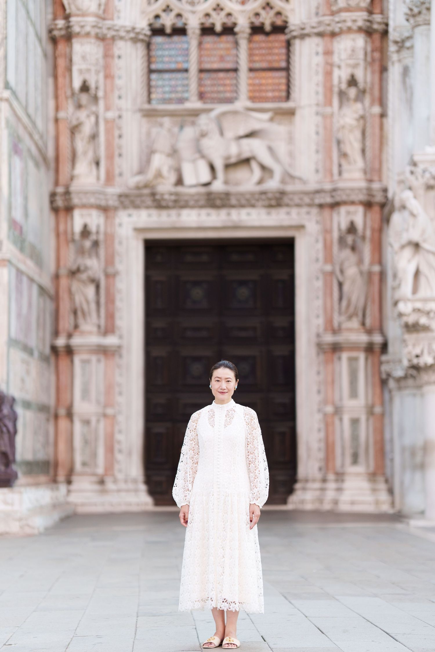 Woman in white lace dress standing before ornate Venice cathedral facade with lion sculpture.