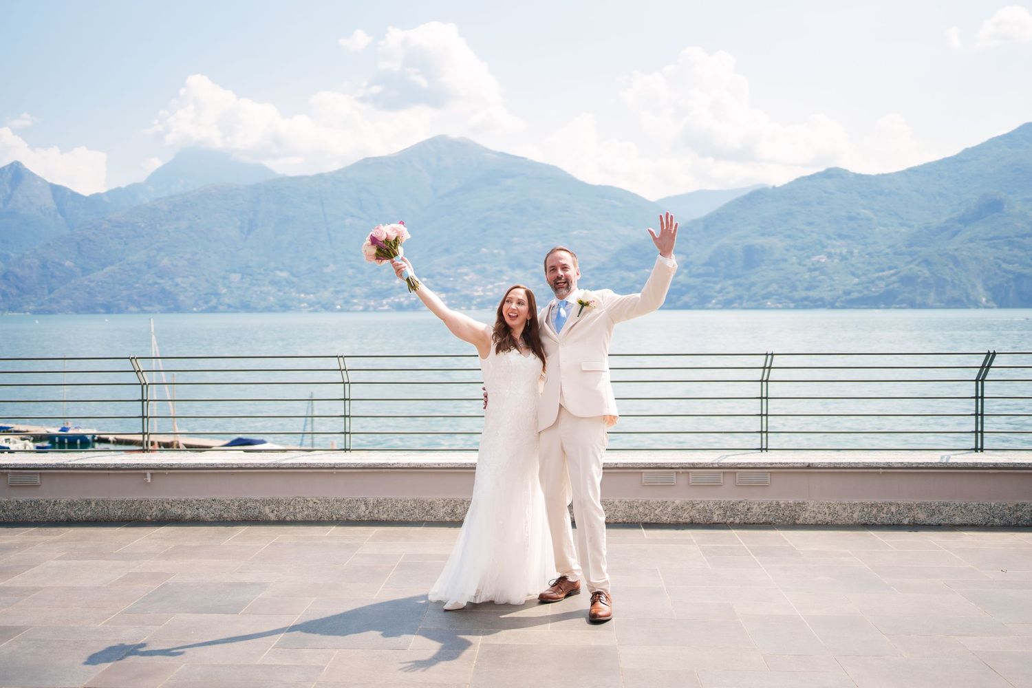 Joyful newlyweds celebrate on a lakeside terrace with mountain backdrop, bride holding bouquet, both raising arms in triumph como lake