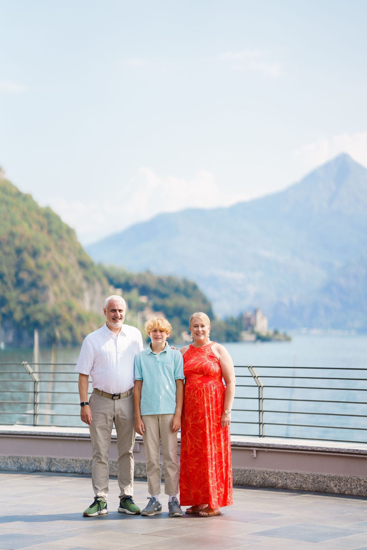 Family of three posing on lakeside terrace with mountain backdrop on sunny day.