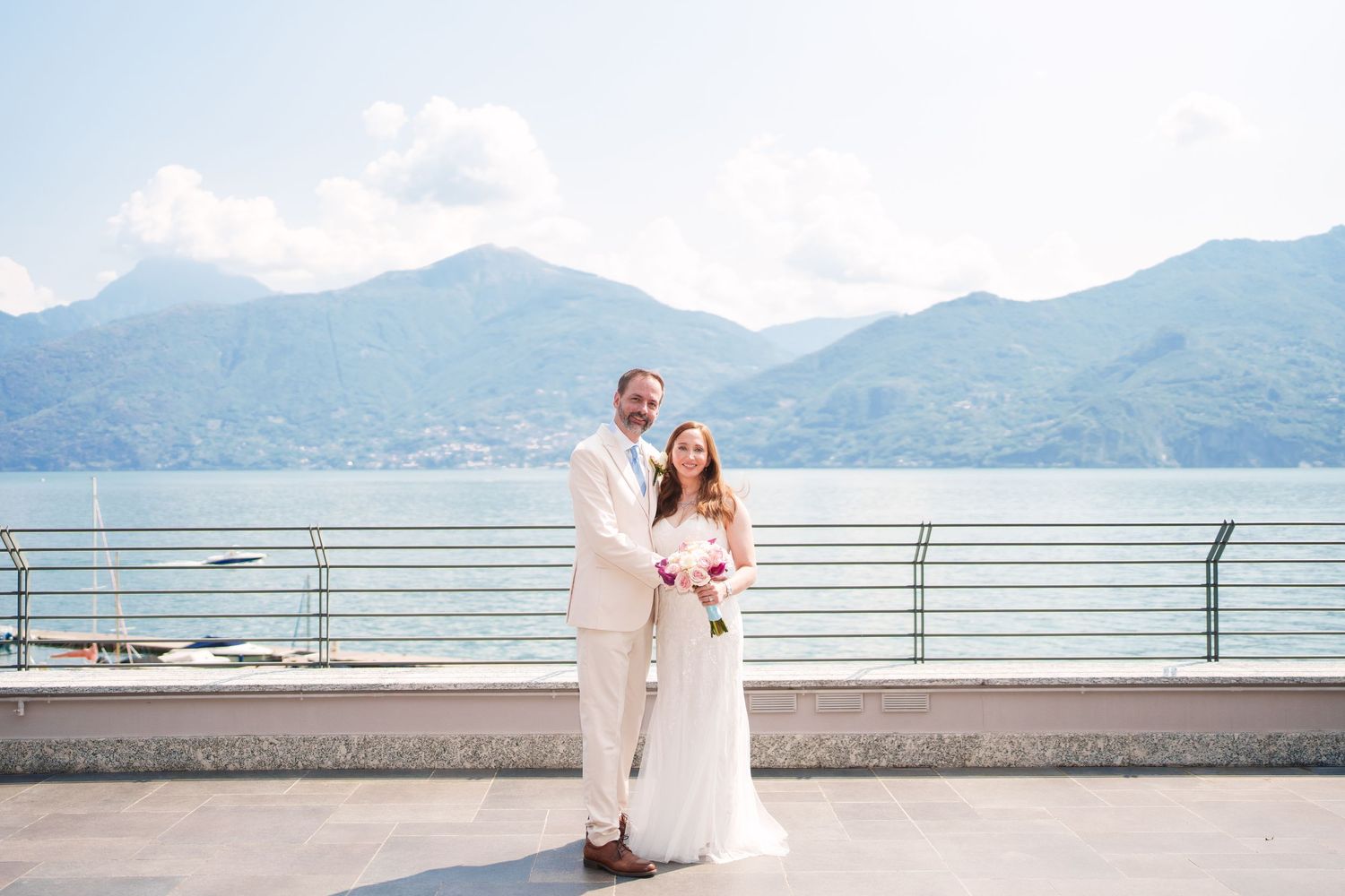 Bride and groom pose on a lakeside terrace with mountain views at their destination wedding in Lake Como, Italy.