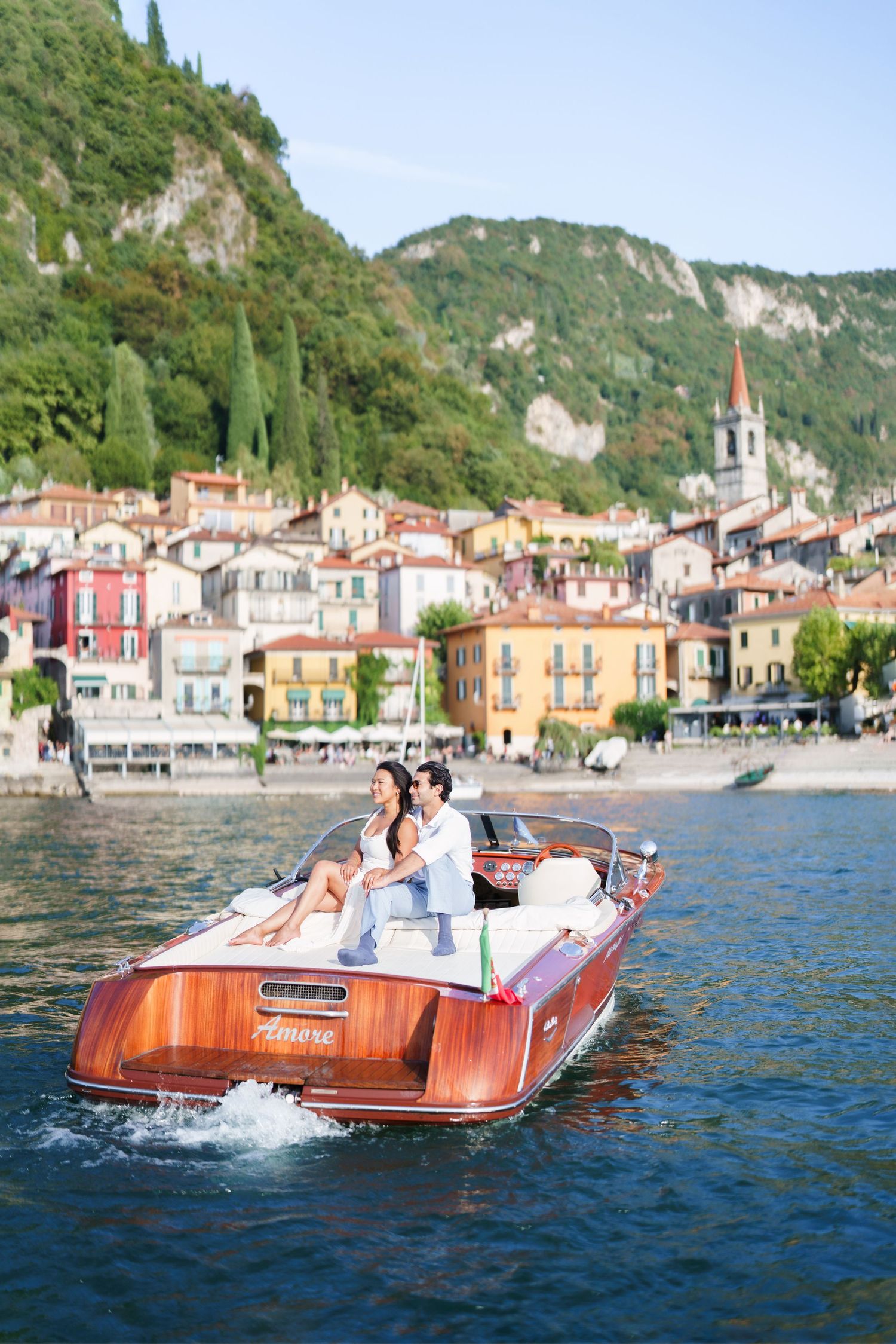 Couple enjoying a romantic boat ride on Lake Como, Italy, with colorful Varenna village and lush green hills in the background.