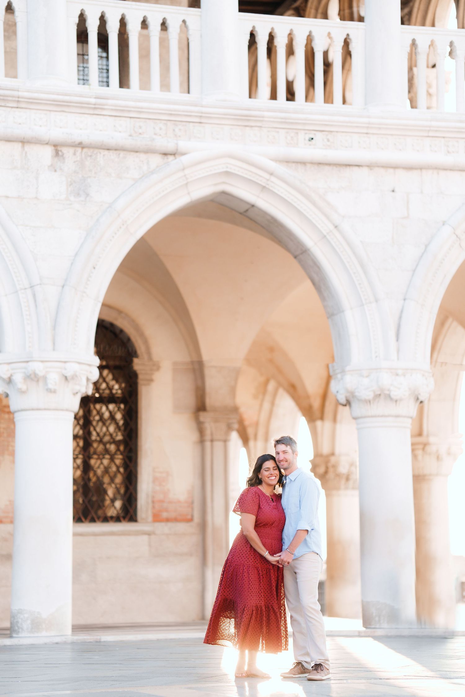 Couple embracing under Gothic arches of Doge's Palace in Venice, woman in red dress, man in light blue shirt.