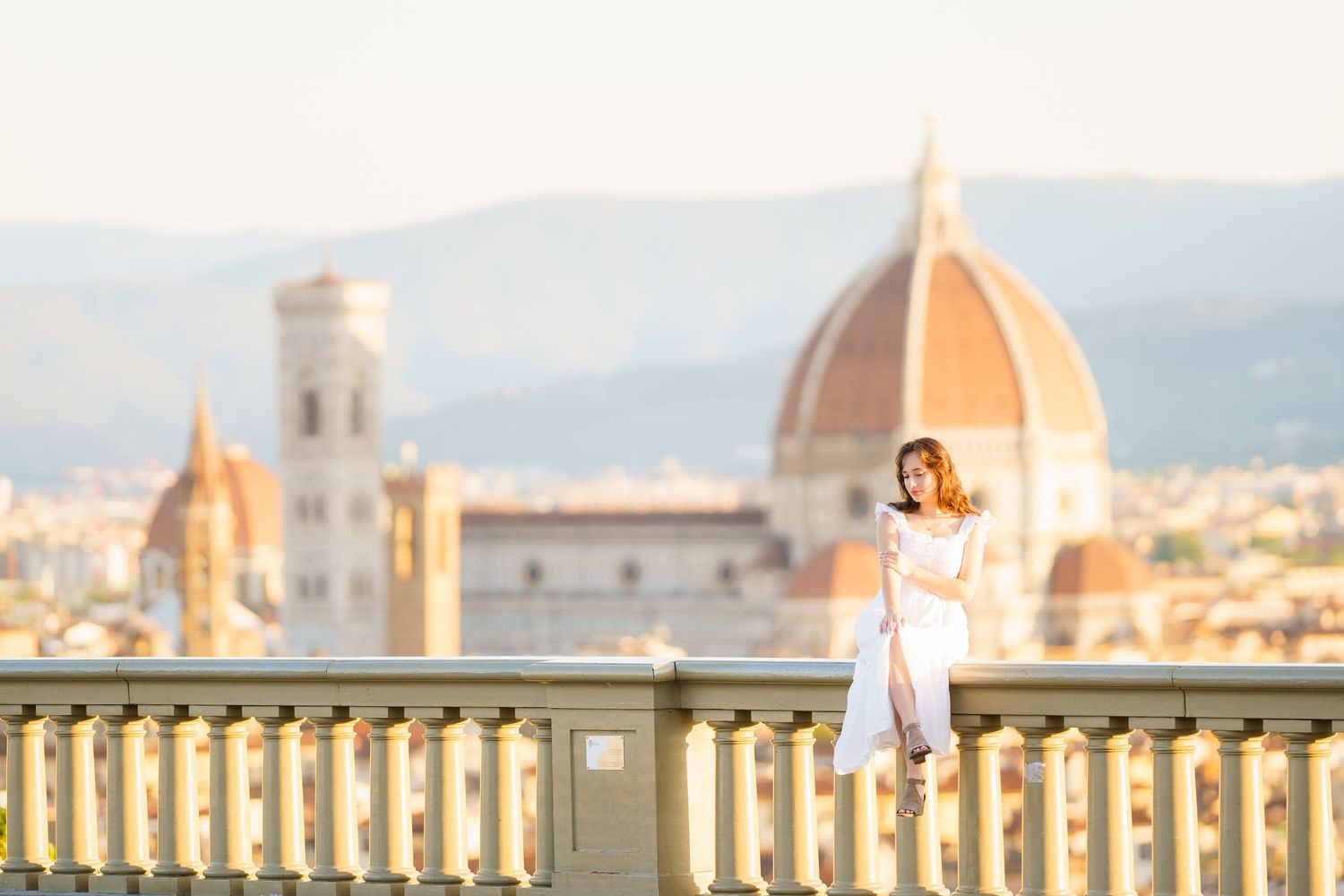 Woman in white dress sitting on a balcony railing with Florence Cathedral dome in the background.