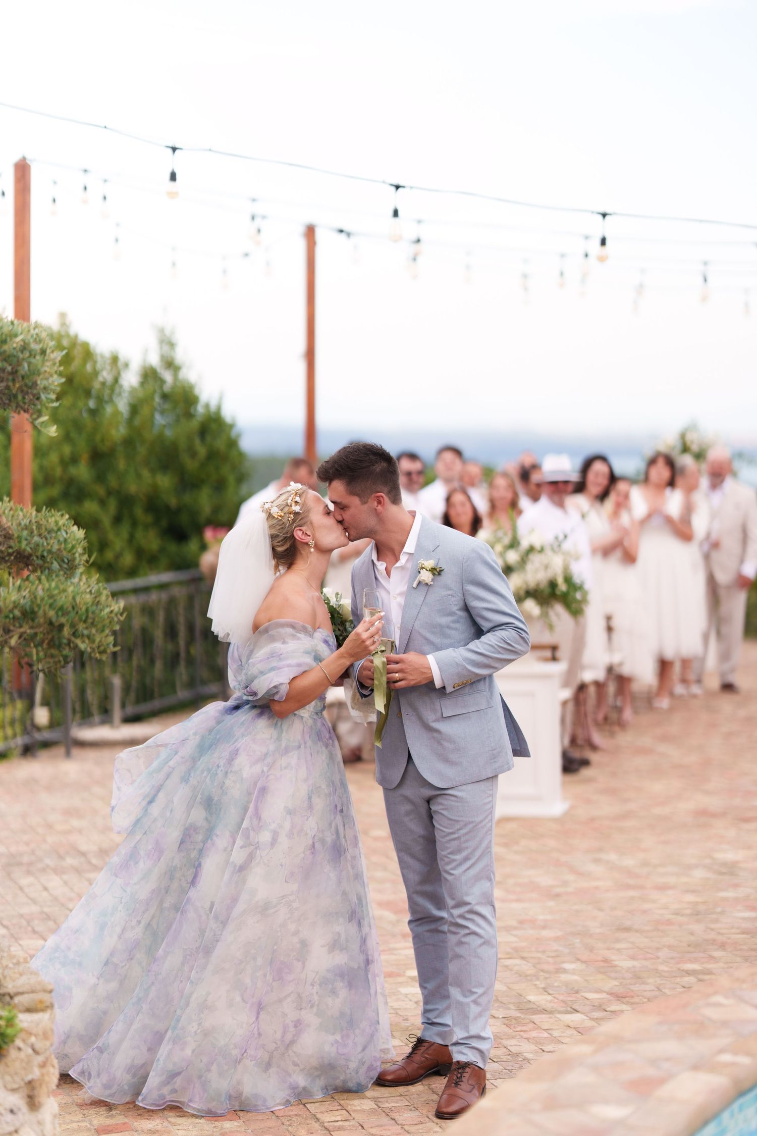 Bride in floral gown and groom in gray suit share first kiss at outdoor wedding ceremony with guests applauding.