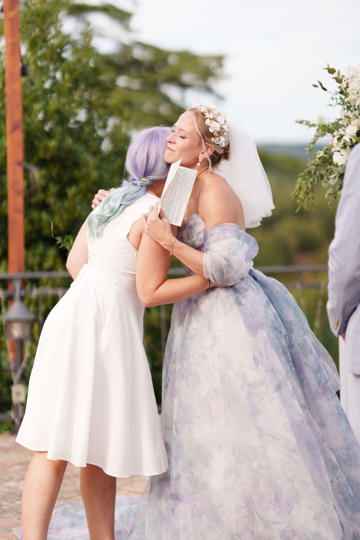 Two brides in white and lavender gowns share a heartfelt hug at an outdoor wedding ceremony.