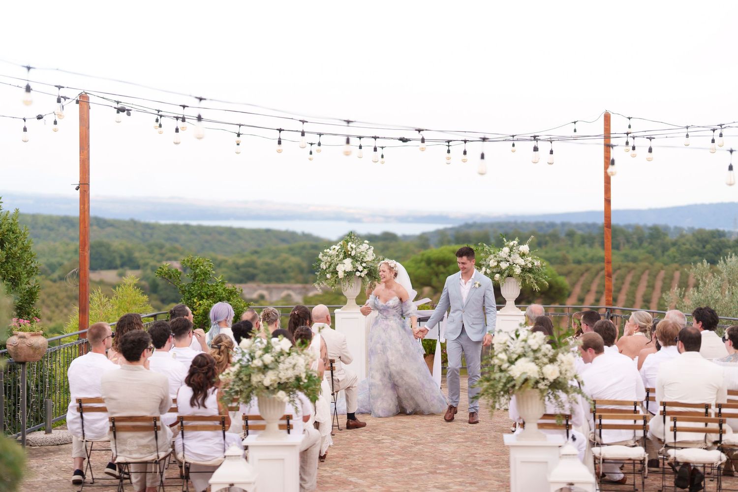 Outdoor vineyard wedding ceremony with couple at altar, floral arrangements, string lights, and scenic landscape backdrop.
