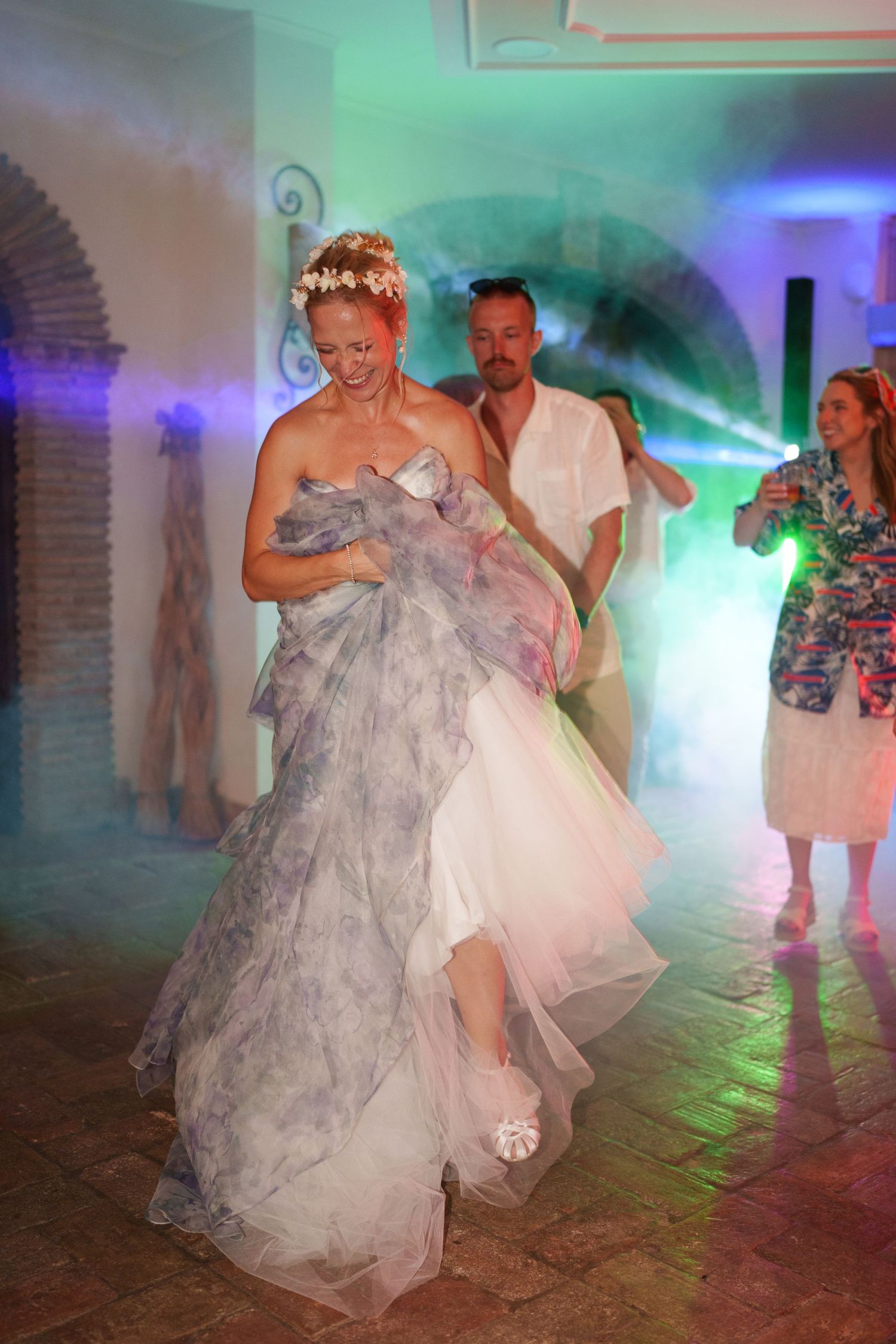 Bride dancing at wedding reception in elegant white and gray gown with floral headpiece under colorful lights.