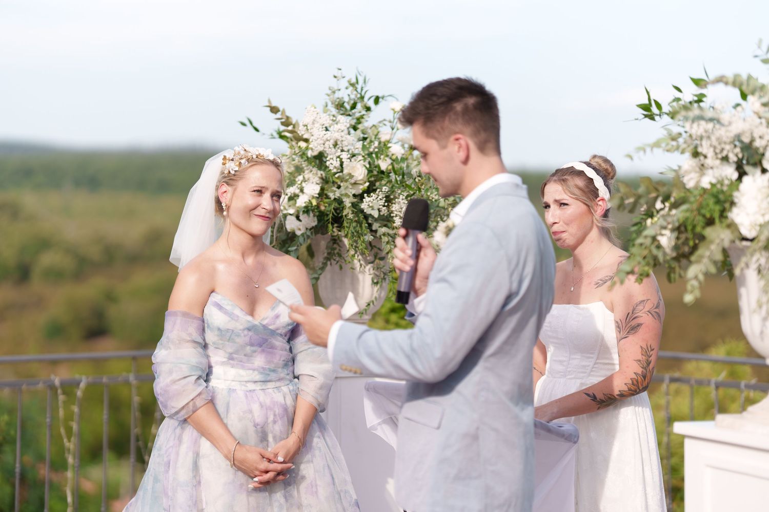 Bride and groom exchange vows outdoors at elegant wedding ceremony with white floral arch and bridesmaid.