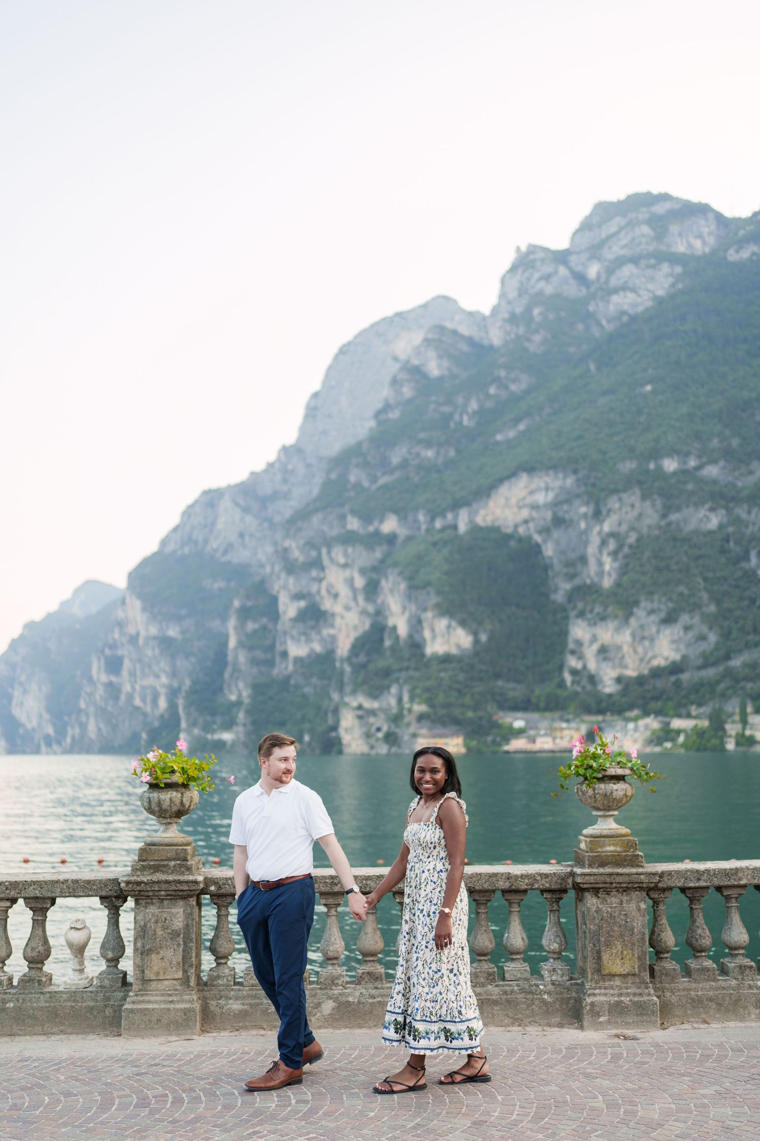 Interracial couple holding hands and smiling on a scenic lakeside terrace with turquoise water and mountains.