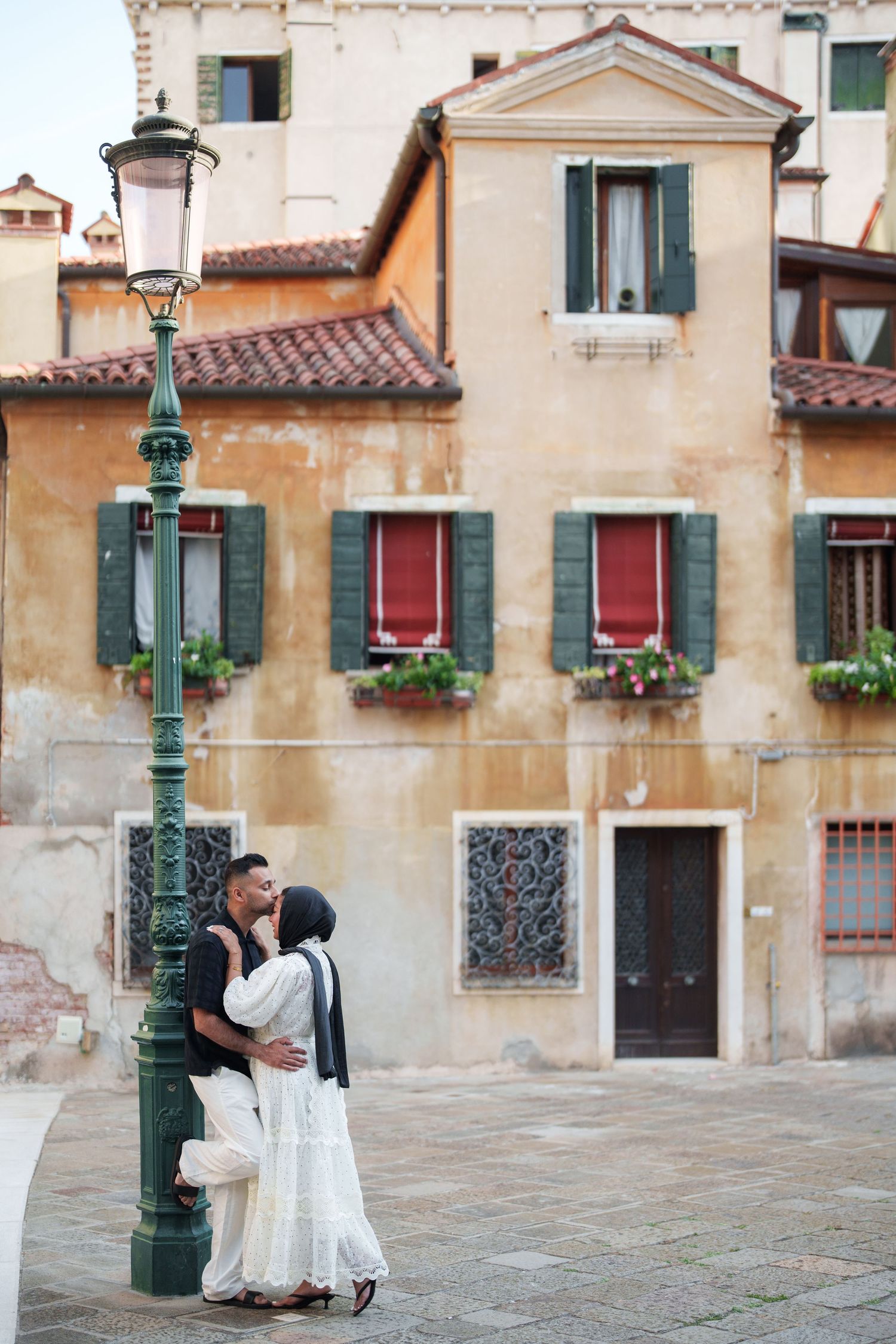 Couple kissing by a vintage lamp post in Venice, Italy, with rustic buildings and green shutters in the background.