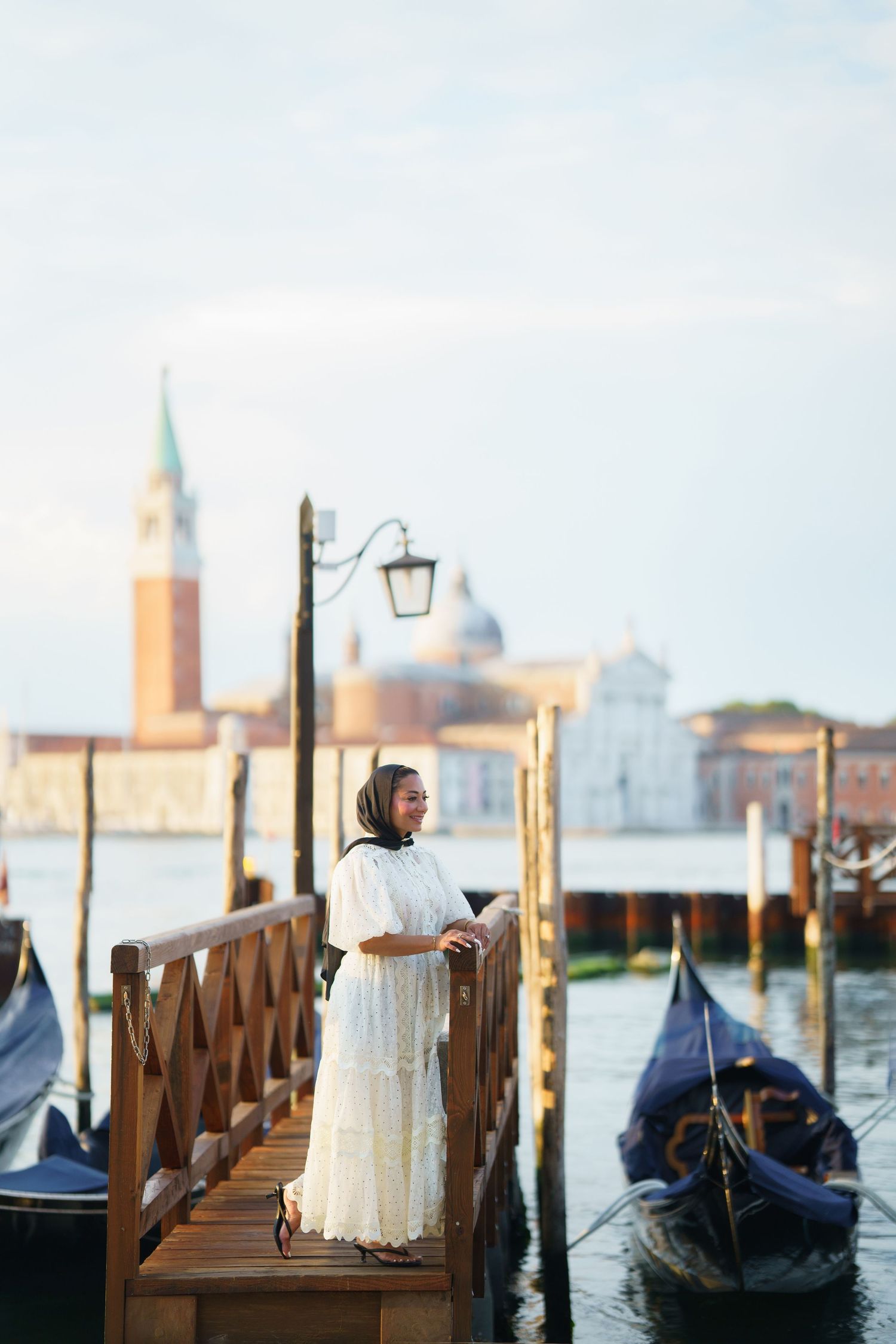 Woman in white dress standing on a wooden dock in Venice, Italy, with gondolas and San Giorgio Maggiore in background.