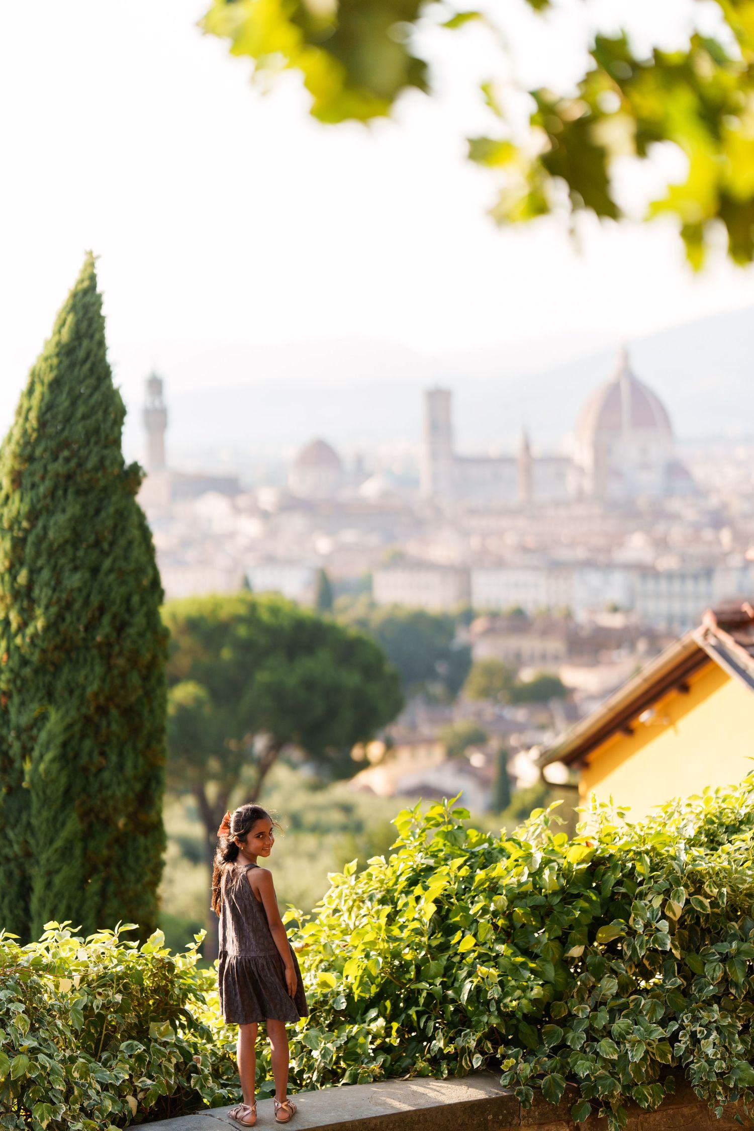Young woman in dress stands on wall overlooking Florence skyline with Duomo cathedral visible in warm sunlight.