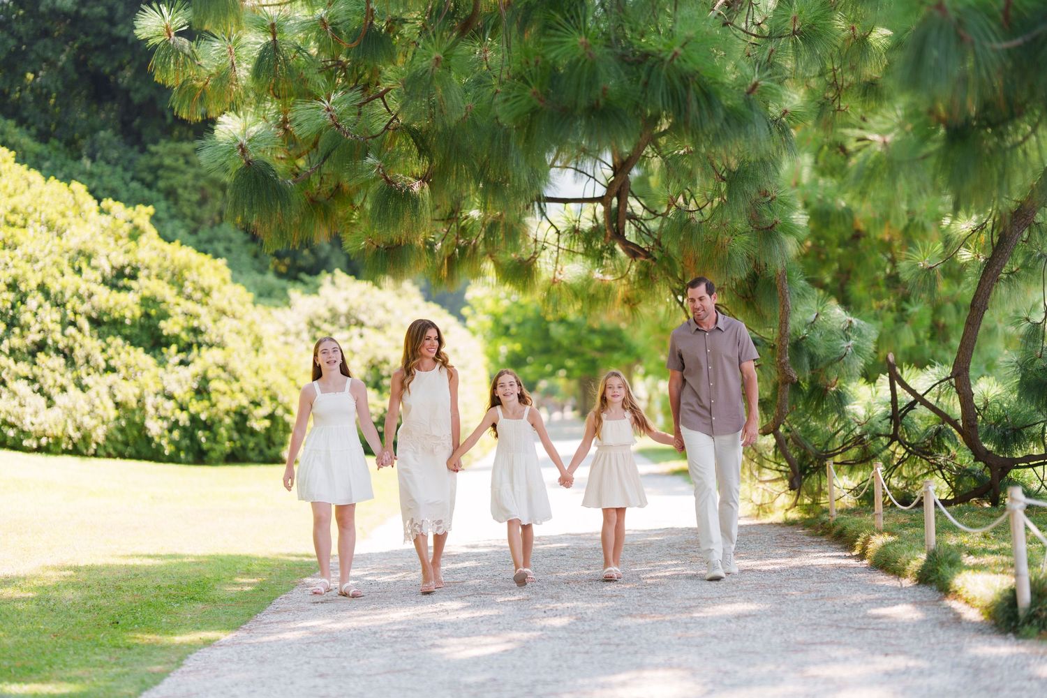Family of five walking hand-in-hand on a garden path surrounded by lush green trees in matching white outfits.