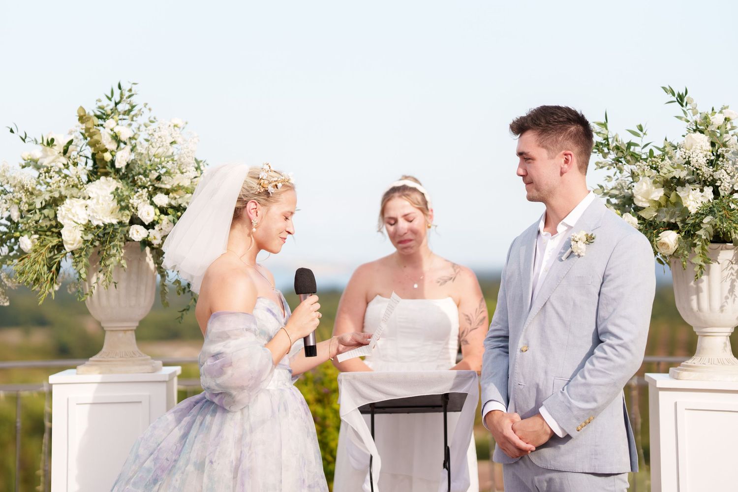 Bride reads wedding vows to groom at outdoor ceremony with white floral arrangements and officiant.