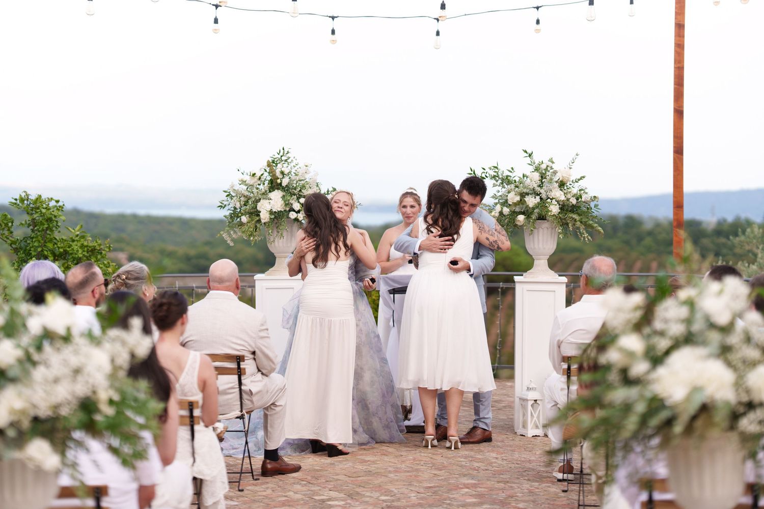 Outdoor wedding ceremony on a terrace with white floral arrangements, guests seated, and scenic mountain views in the background.