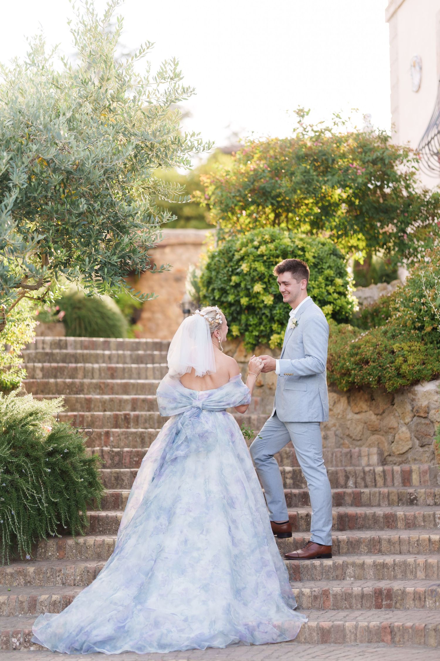 Bride in blue floral gown and groom in gray suit hold hands on brick stairs surrounded by lush greenery.