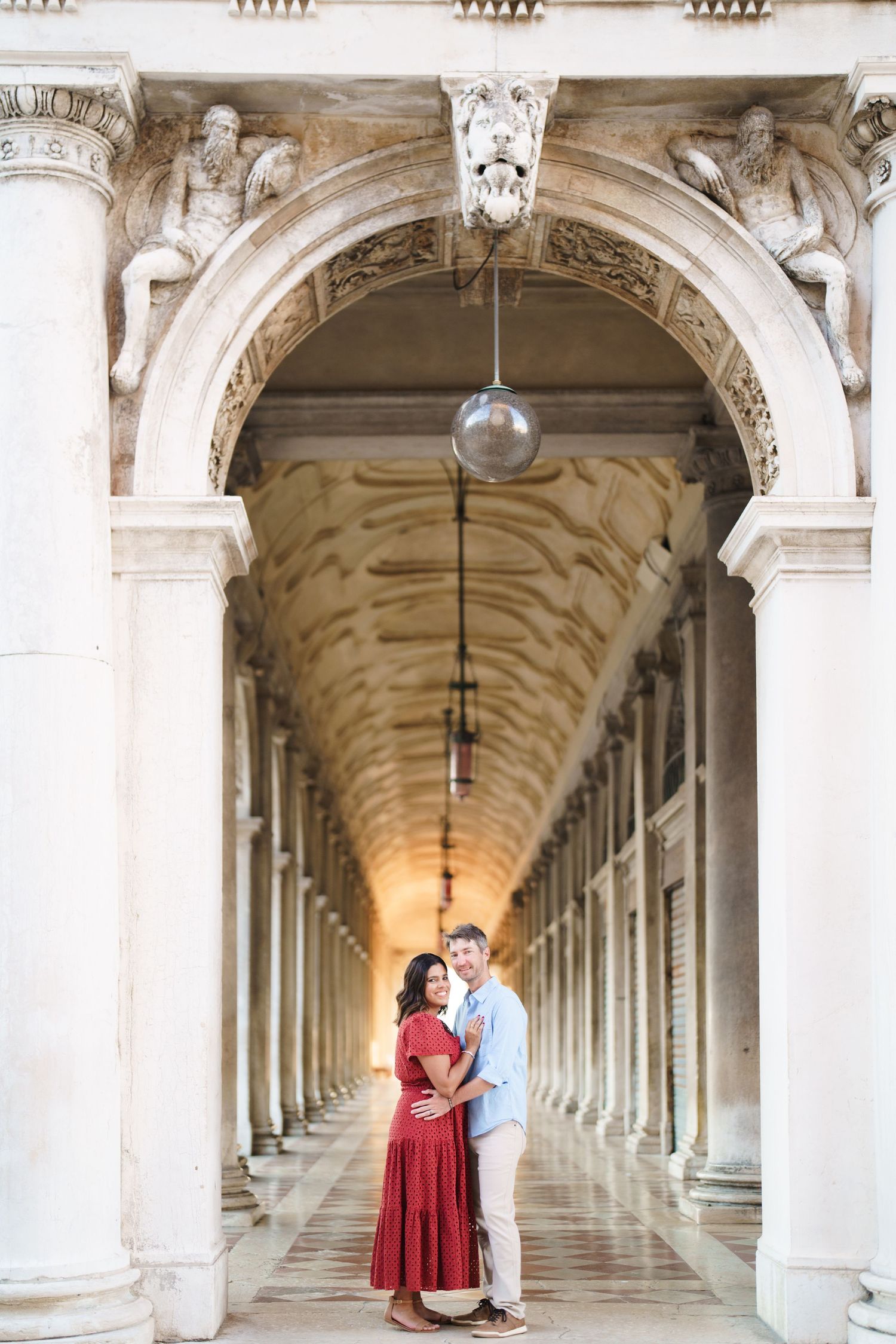 Couple embracing under ornate marble archway colonnade in Venice, woman in red dress, man in light blue shirt.